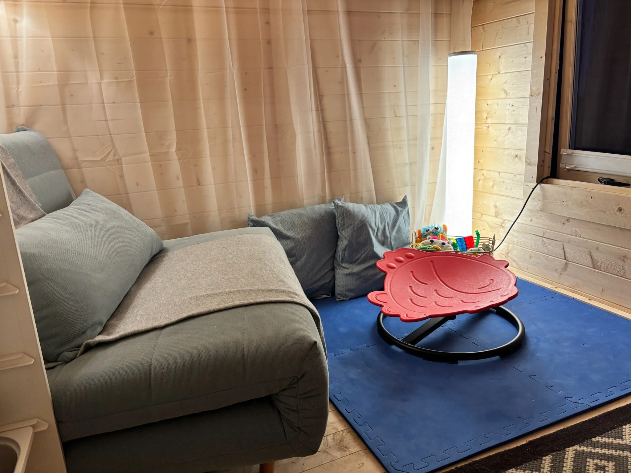 A cozy room with a light gray sofa with several pillows, a blue foam puzzle mat on the wooden floor, a small red table, a toy basket filled with plush toys, a bright window with a roller shade, and a corner of a black electronic device.