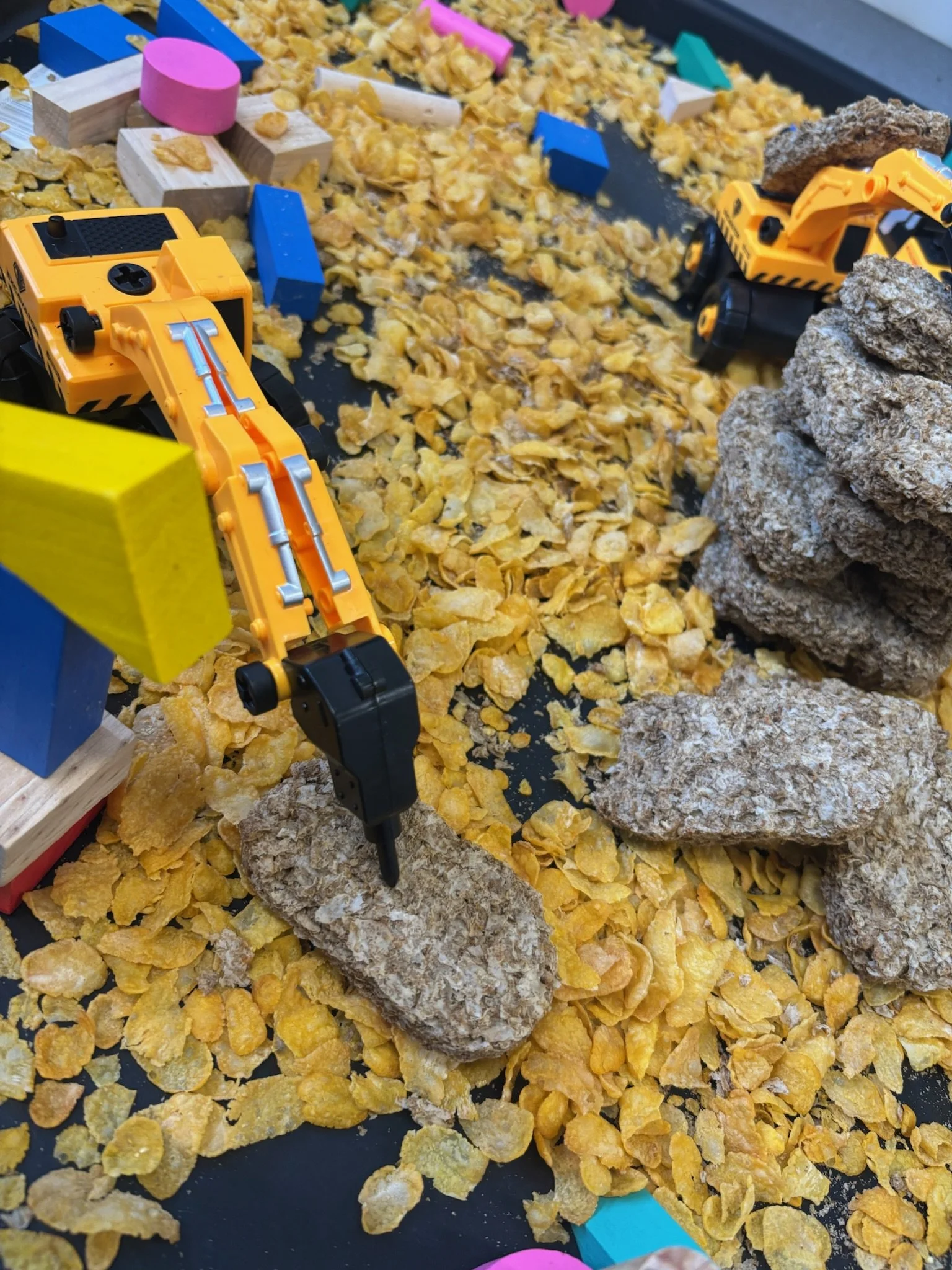 A model landscape with yellow-orange crushed cereal or flakes, large rocks, blue and pink blocks, and yellow toy construction vehicles on a black surface.