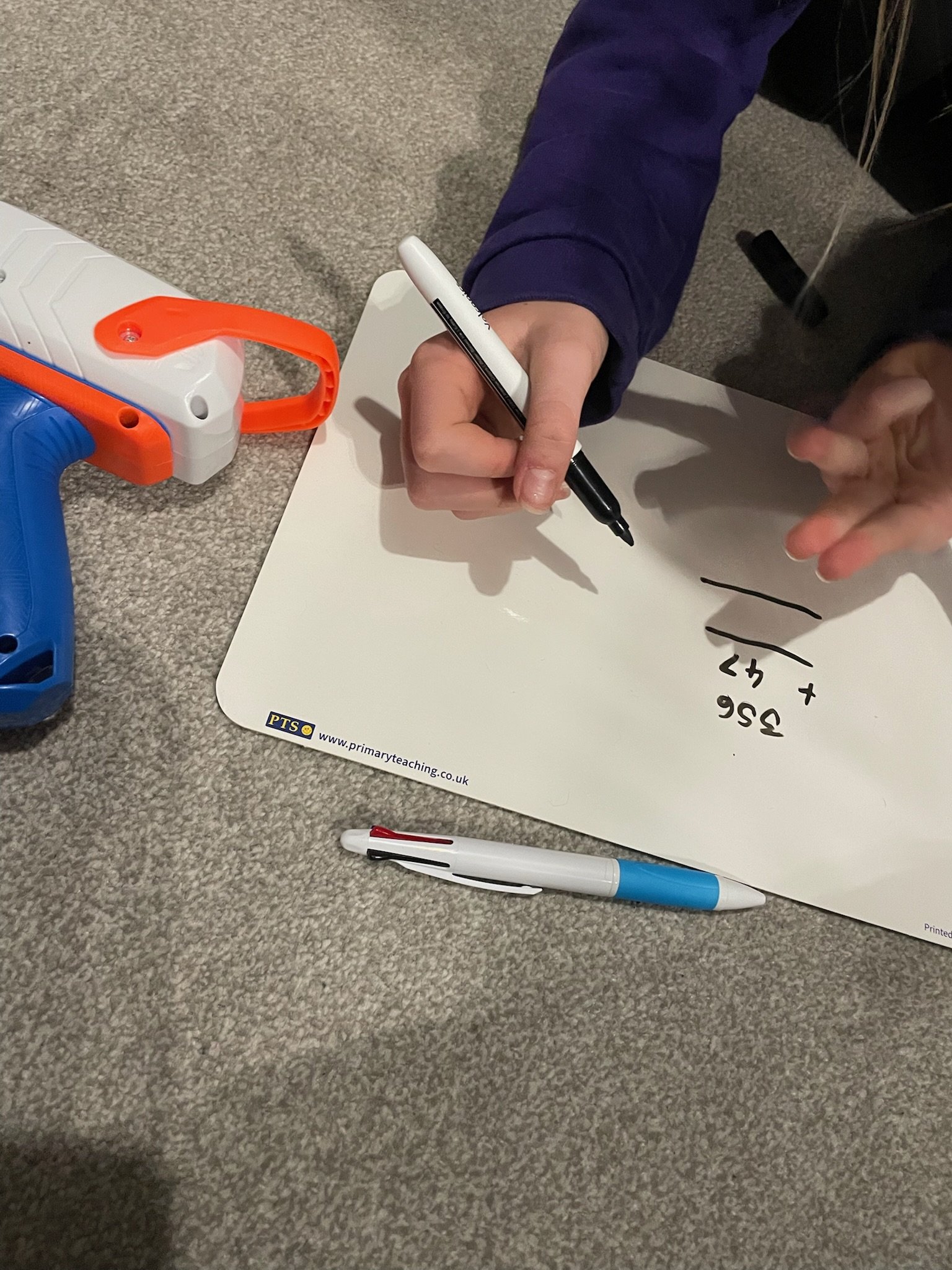 A person wearing a blue jacket is writing math equations on a whiteboard with a black marker. There are various markers, a toy gun, and a piece of paper on a carpeted floor.