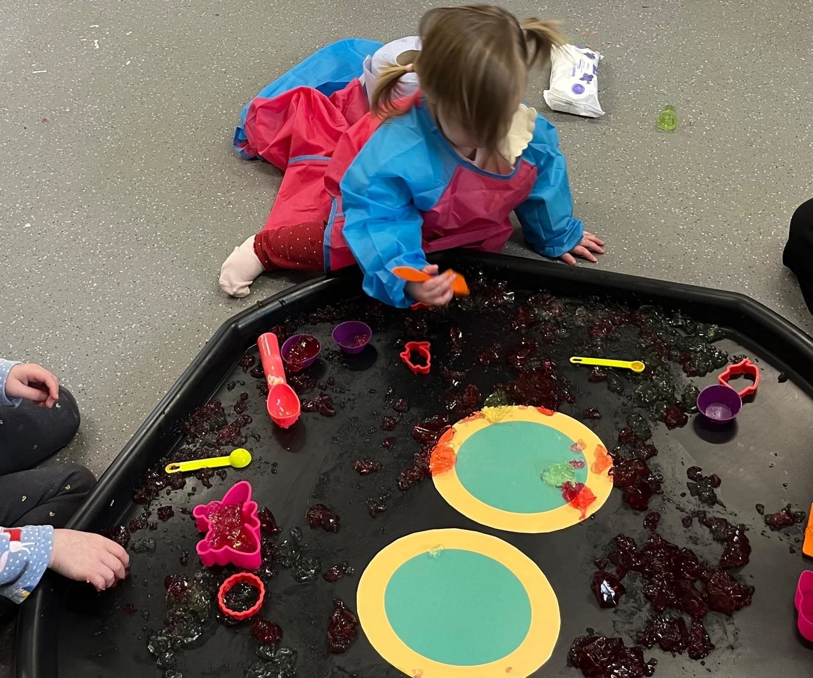 Child in a colorful smock playing with red slime and toys on a black tray with paper plates and bowls nearby in an indoor play area.