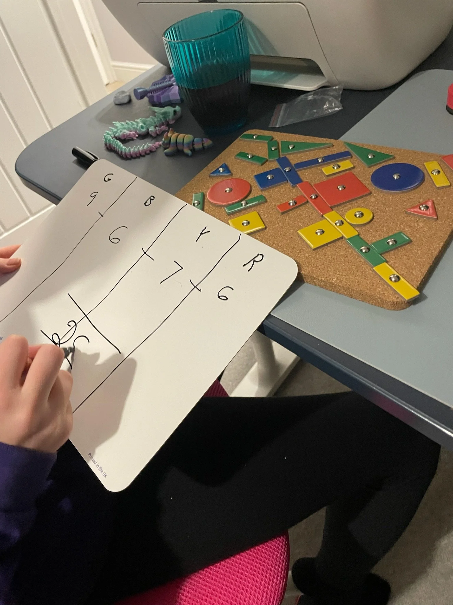 A child holding a whiteboard with a table of numbers and symbols, on a black table with colorful toys, a teal glass, and a sewing pin board, possibly engaged in a learning activity.