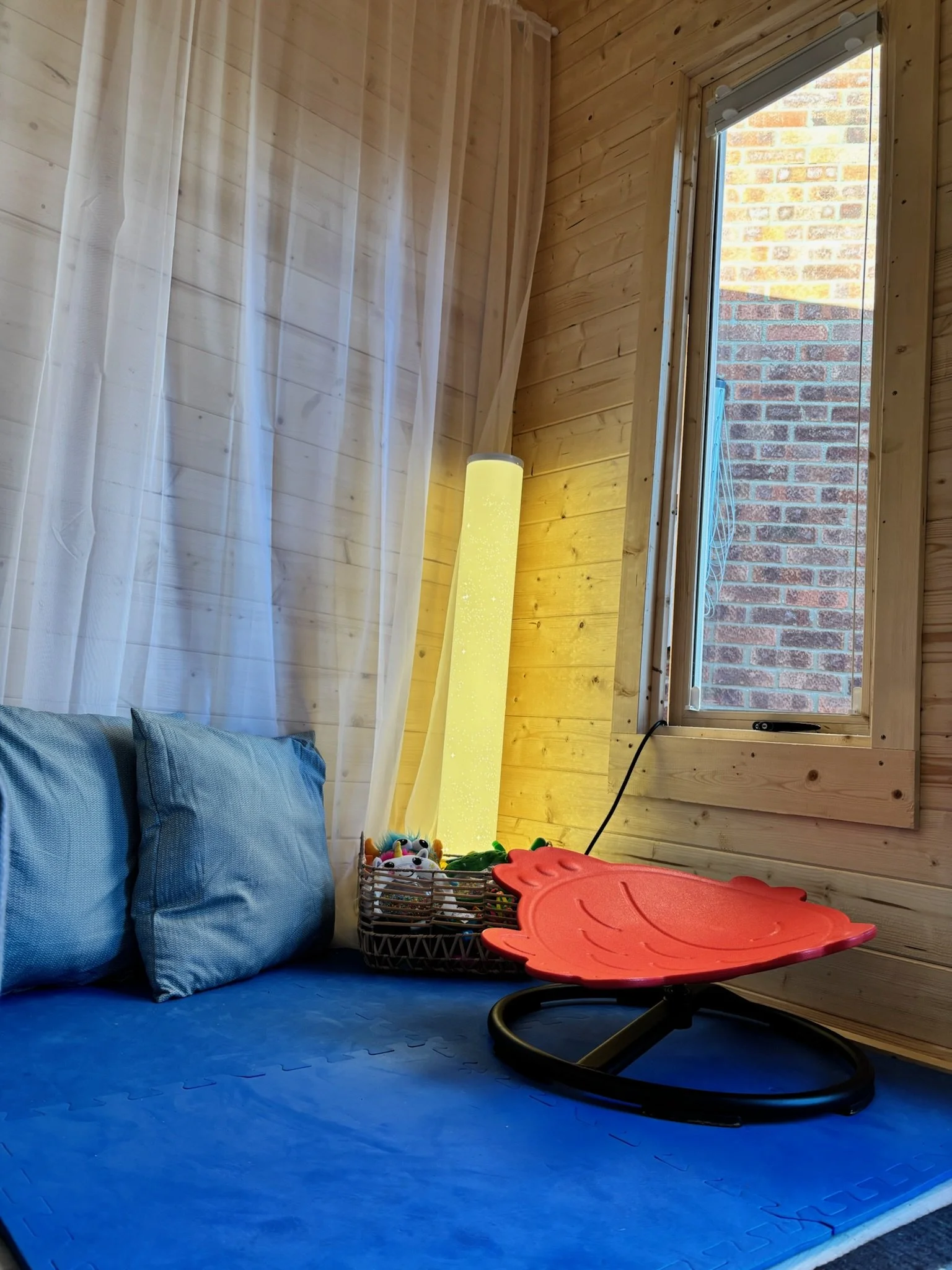 A cozy room corner with light wood-paneled walls, a window showing a brick wall outside, a blue cushioned seating area with two pillows, a basket of children's toys, and a pink rocking chair.