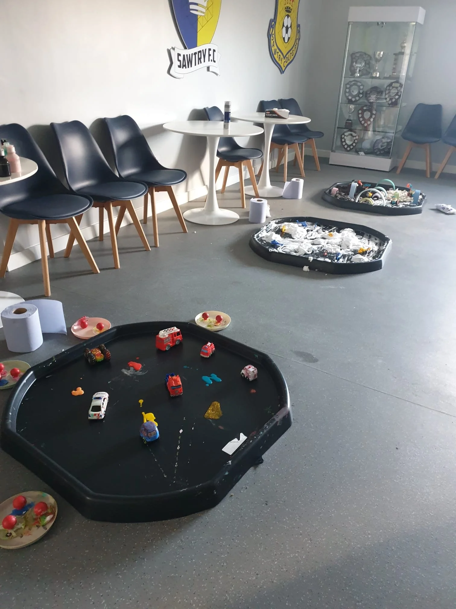 Indoor room with black chairs lined up against a white wall, with a white round table in the middle and a glass display case filled with trophies. The floor has scattered toys, disposable cups, and paper towel rolls. On the wall, there are two sports