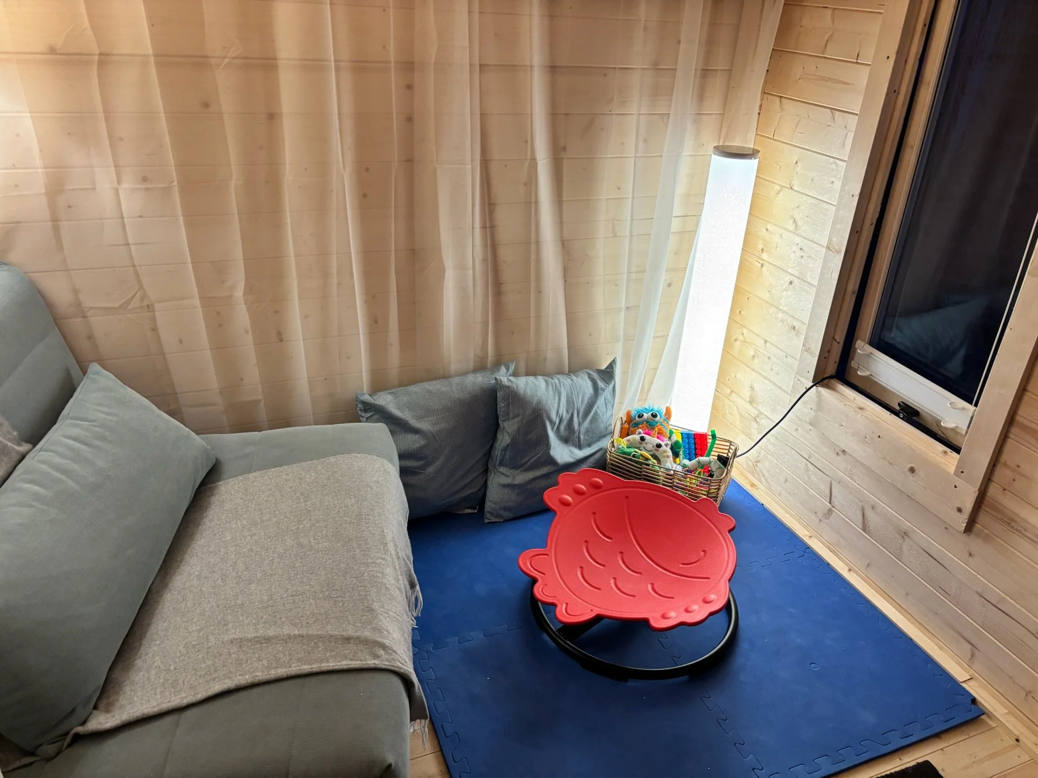 A cozy indoor space with a gray sofa, gray pillows, and a small colorful children's toy basket on a wooden floor with foam mats. A flat-screen TV is on a wooden stand. The room has wooden walls and a ceiling with a hanging light.