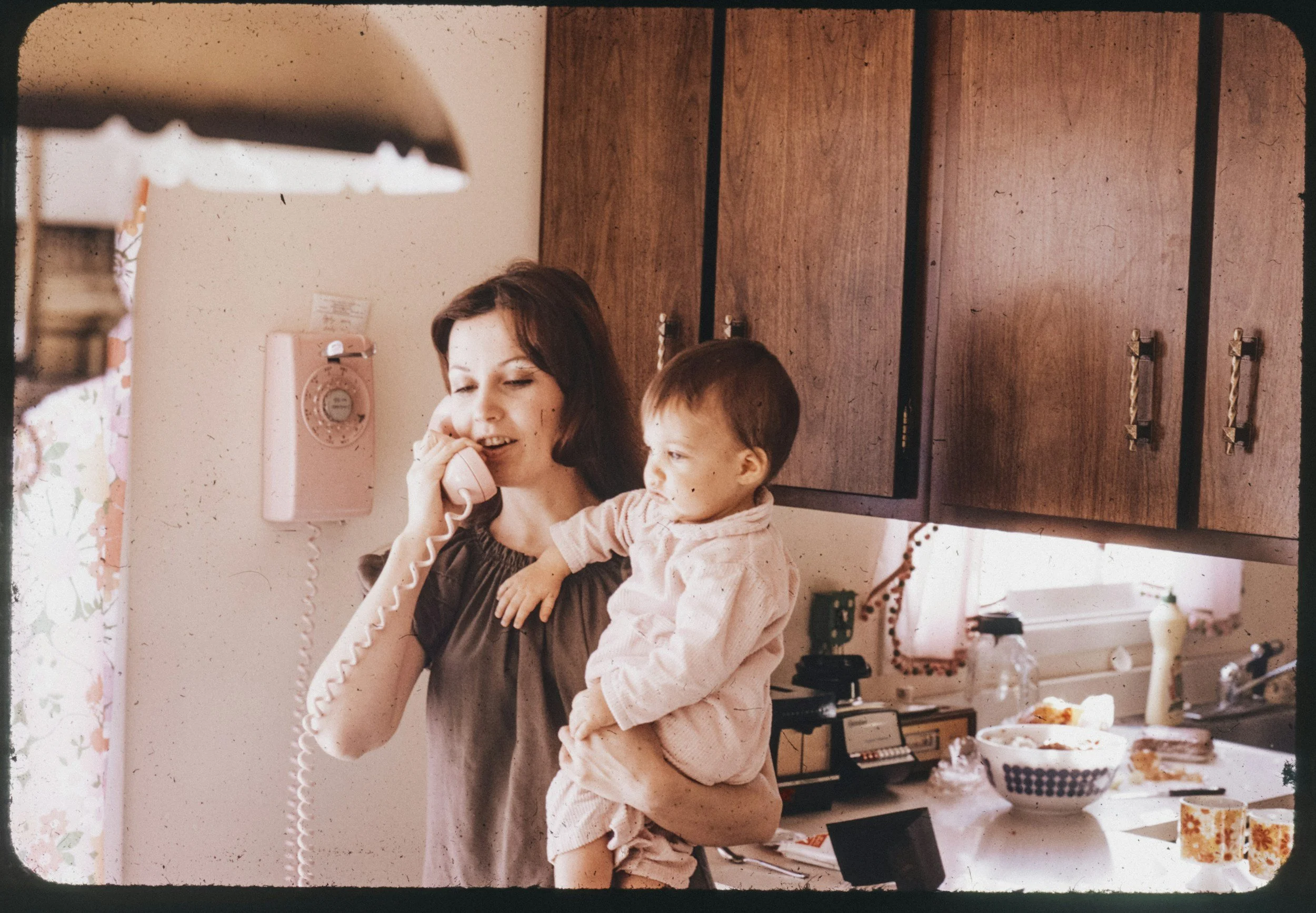 A woman with dark hair talking on a pink rotary phone while holding a young girl with short hair. They are in a kitchen with wooden cabinets, some dishes on the table, and a pink vintage telephone on the wall.
