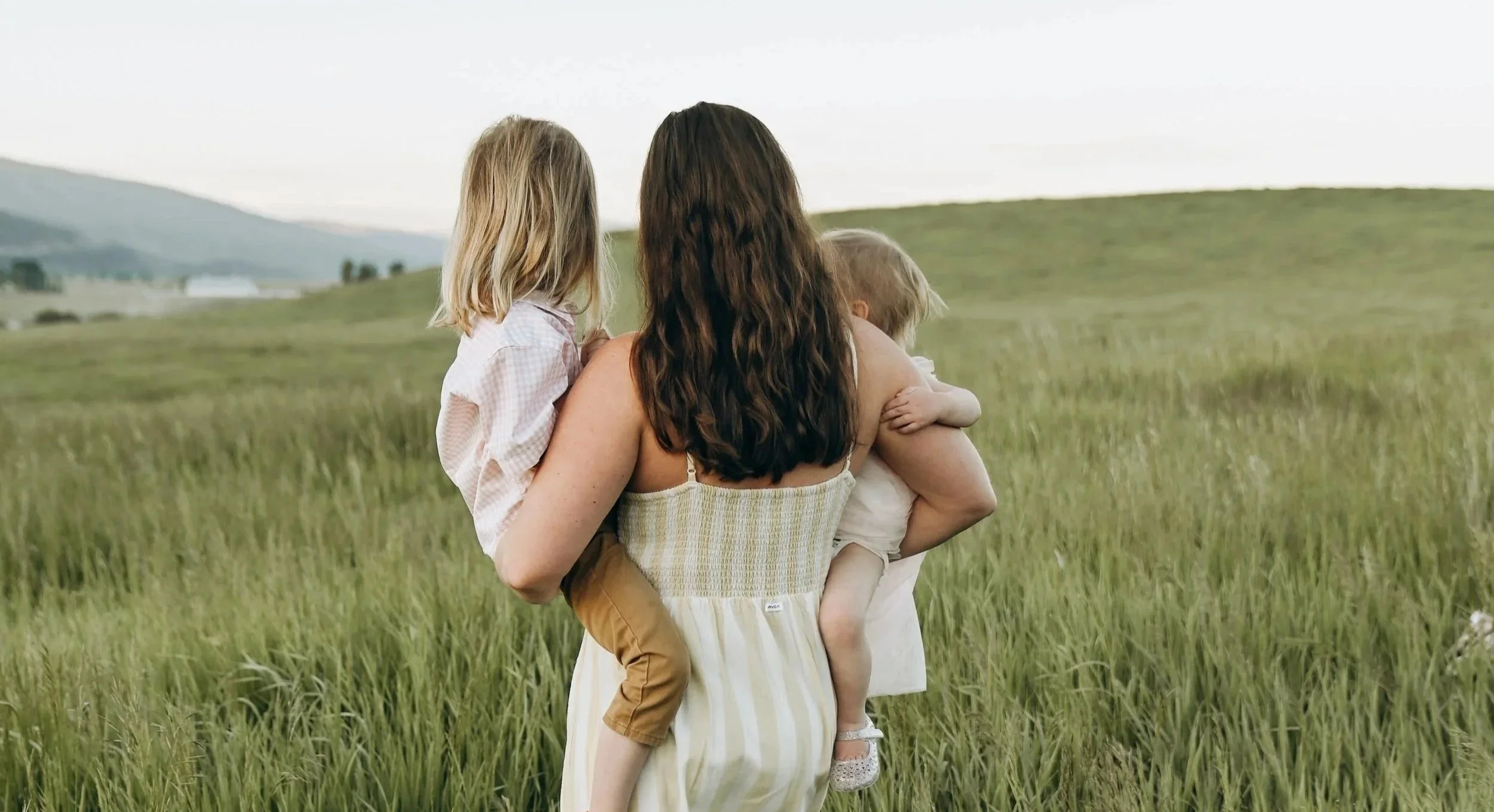 A woman holding two children in a grassy field, with mountains and cloudy sky in the background.