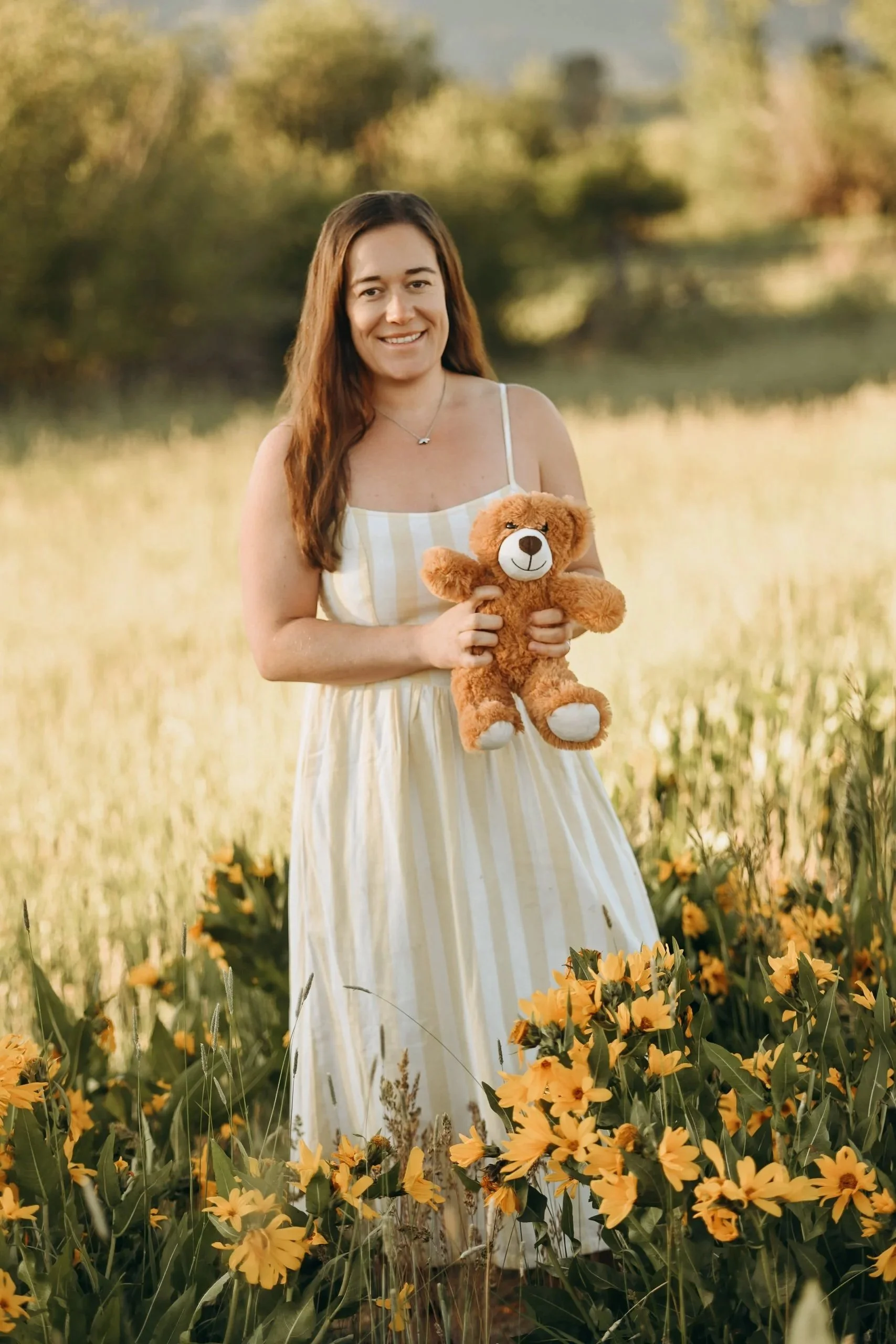 A woman with long brown hair in a white and yellow striped dress holding a teddy bear standing in a field of yellow flowers with trees in the background.