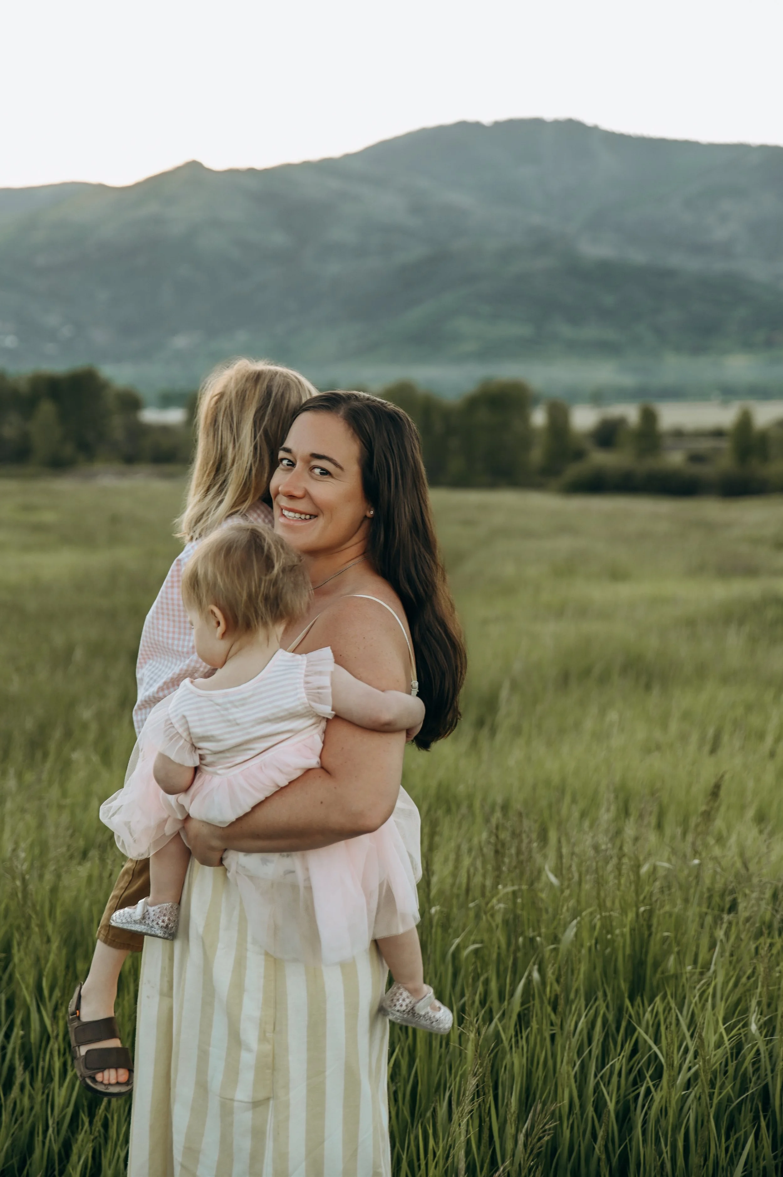 A woman holding two children in a grassy field with mountains in the background.