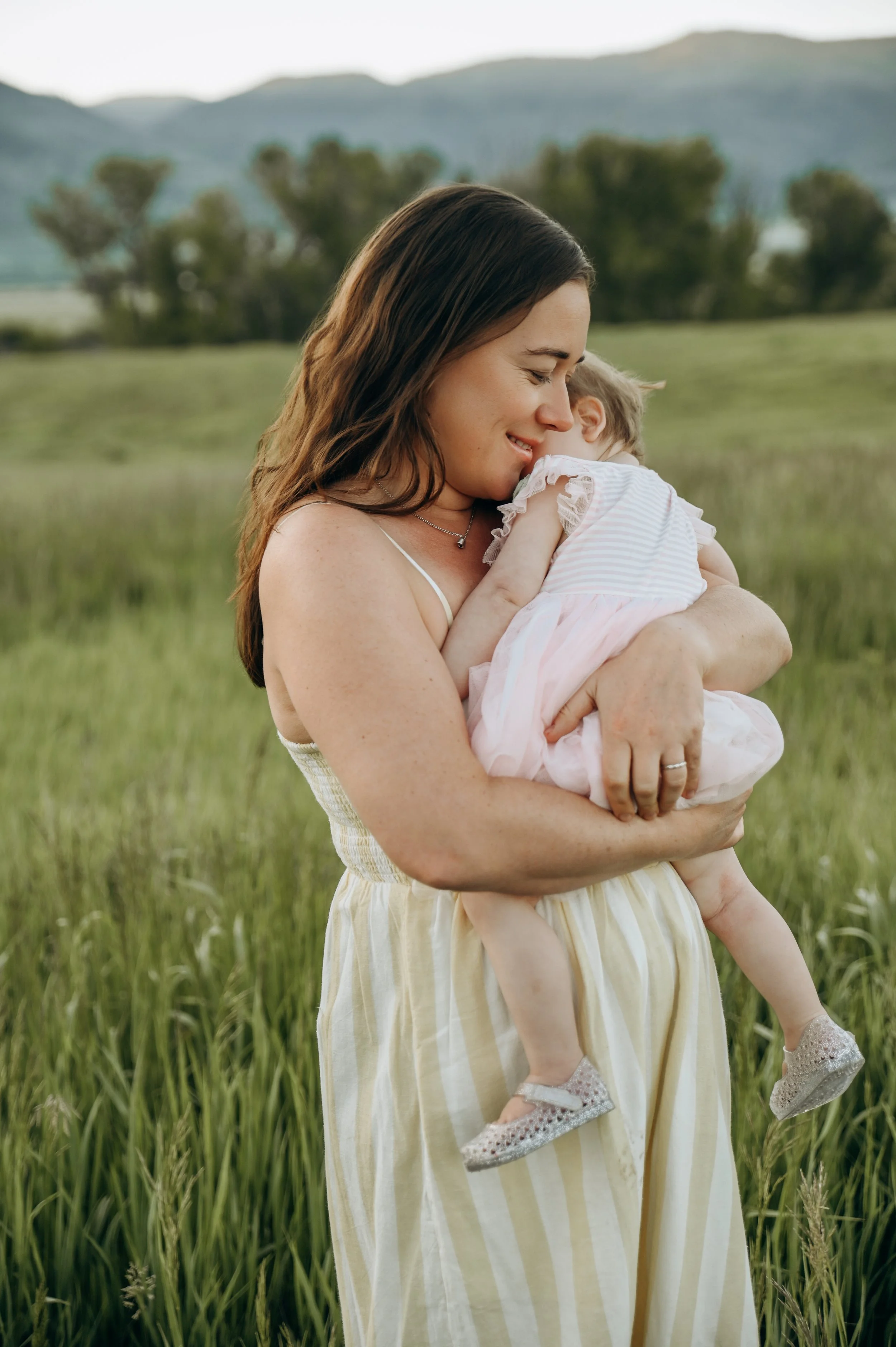 A woman with long brown hair, wearing a yellow and white striped dress, holding a small child in a field of tall grass with trees and mountains in the background.