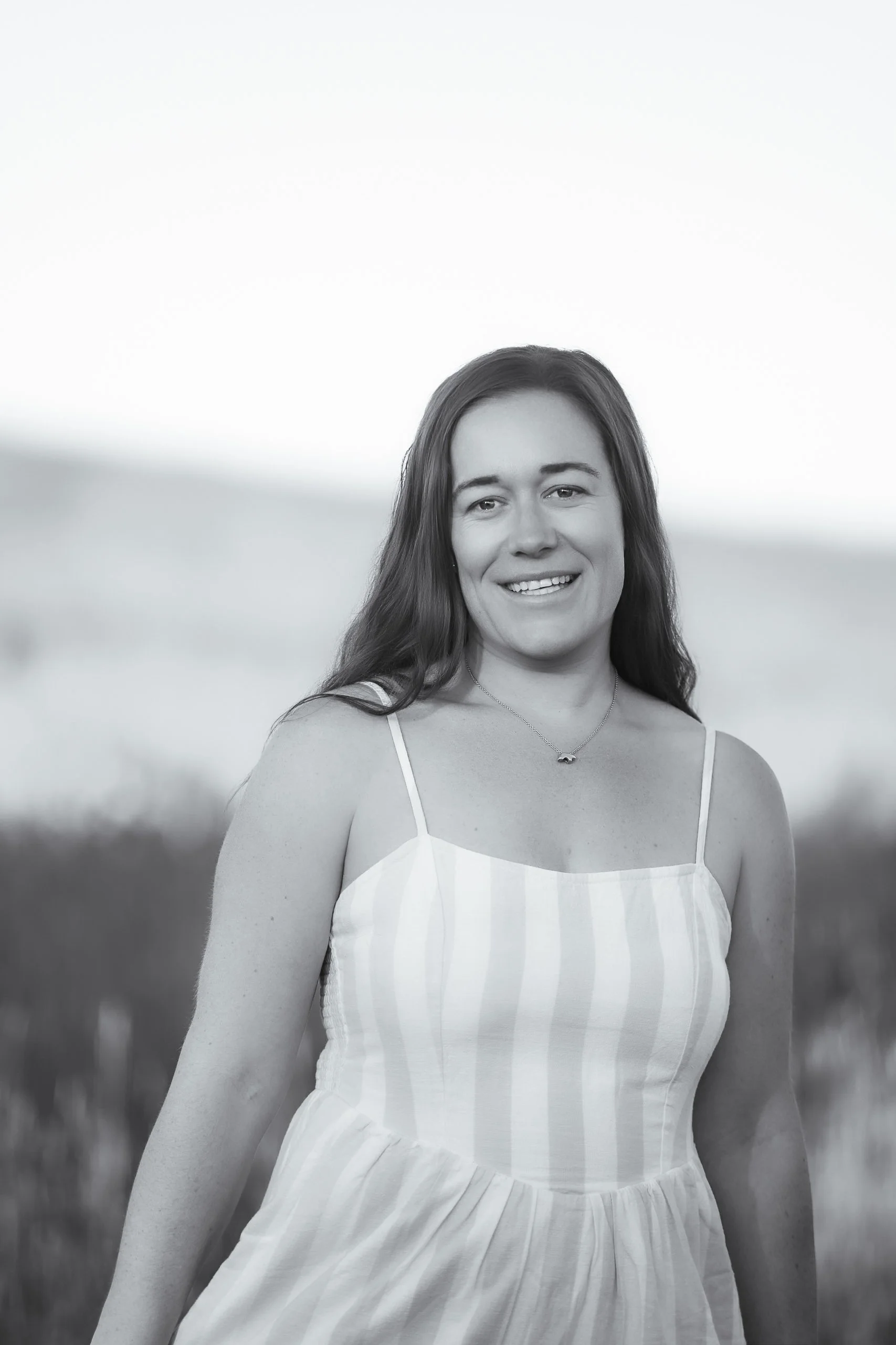 Black and white photo of a young woman with long hair, smiling, wearing a sleeveless striped dress, outdoors with blurred background.
