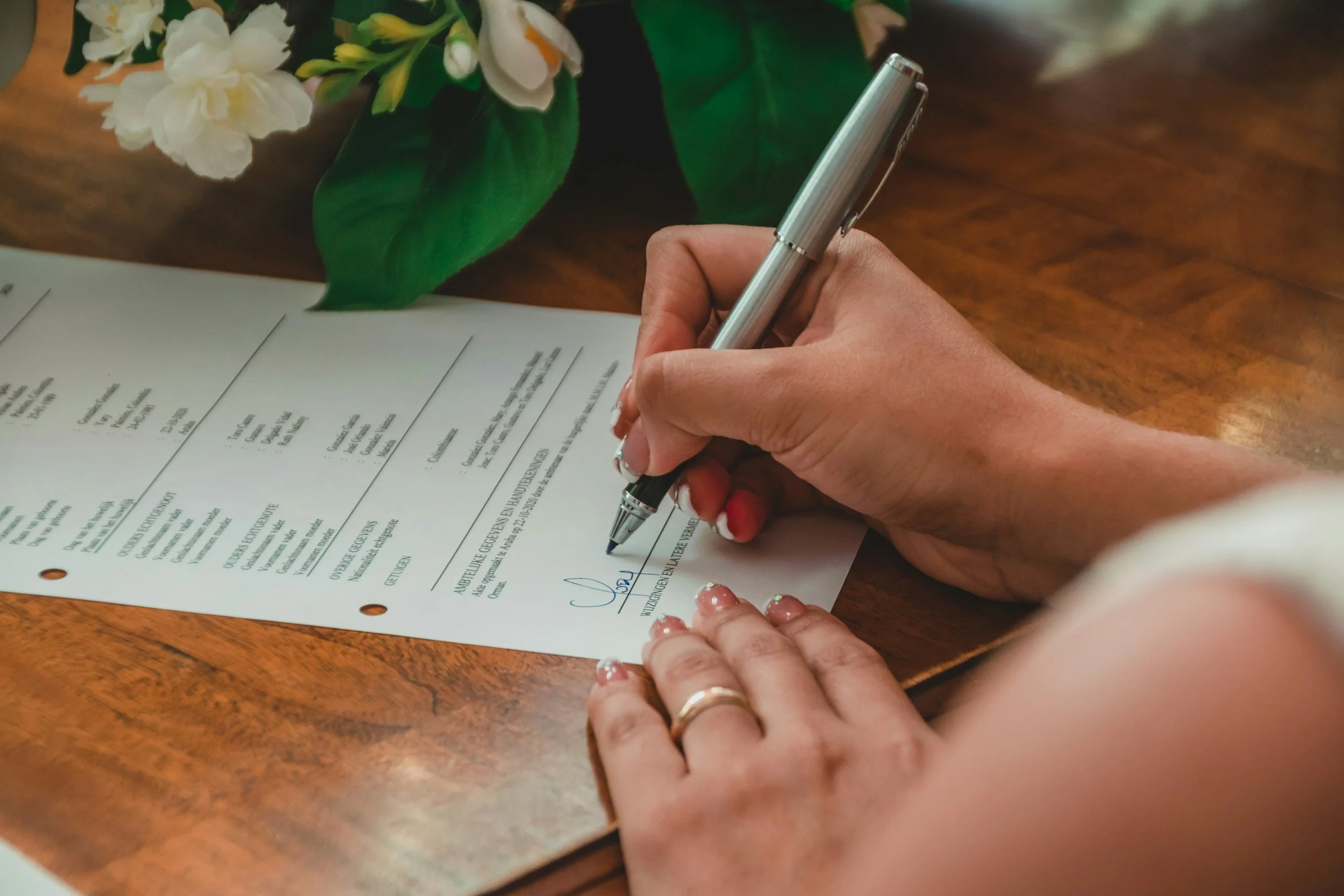 A person signing a document on a wooden table, with a bouquet of white flowers nearby.