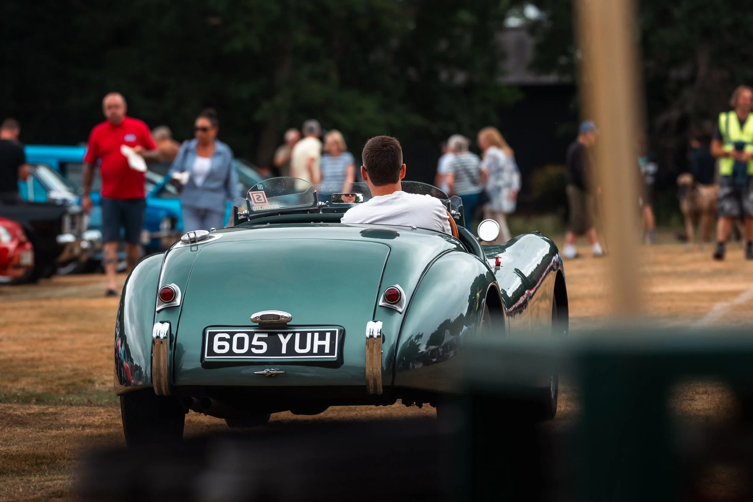 A vintage green sports car with the license plate '605 YUH' parked on a grassy area at an outdoor event with several people in the background, some walking and others standing around, with trees surrounding the scene.