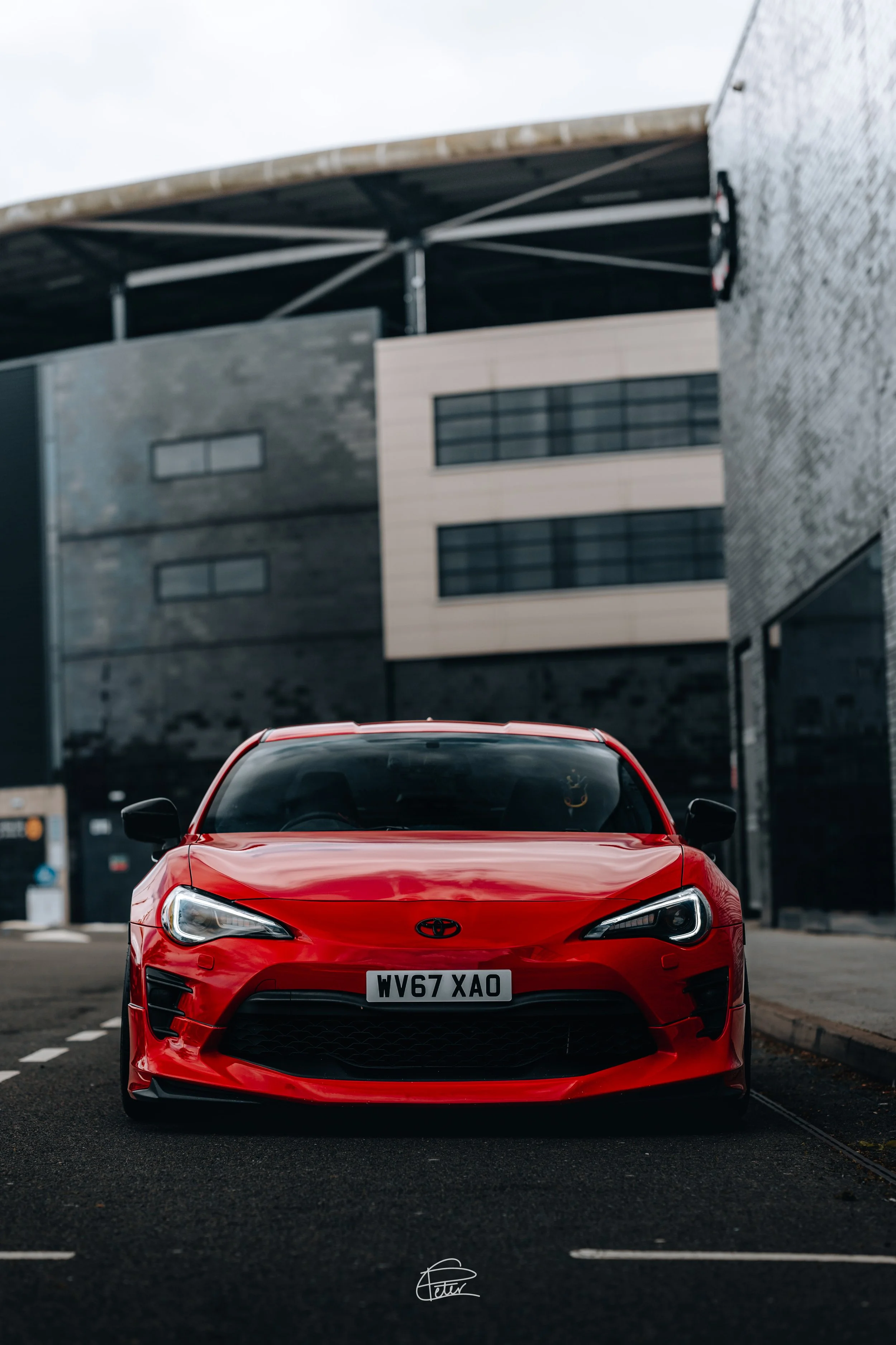 Red Toyota car parked on city street with modern buildings in background.