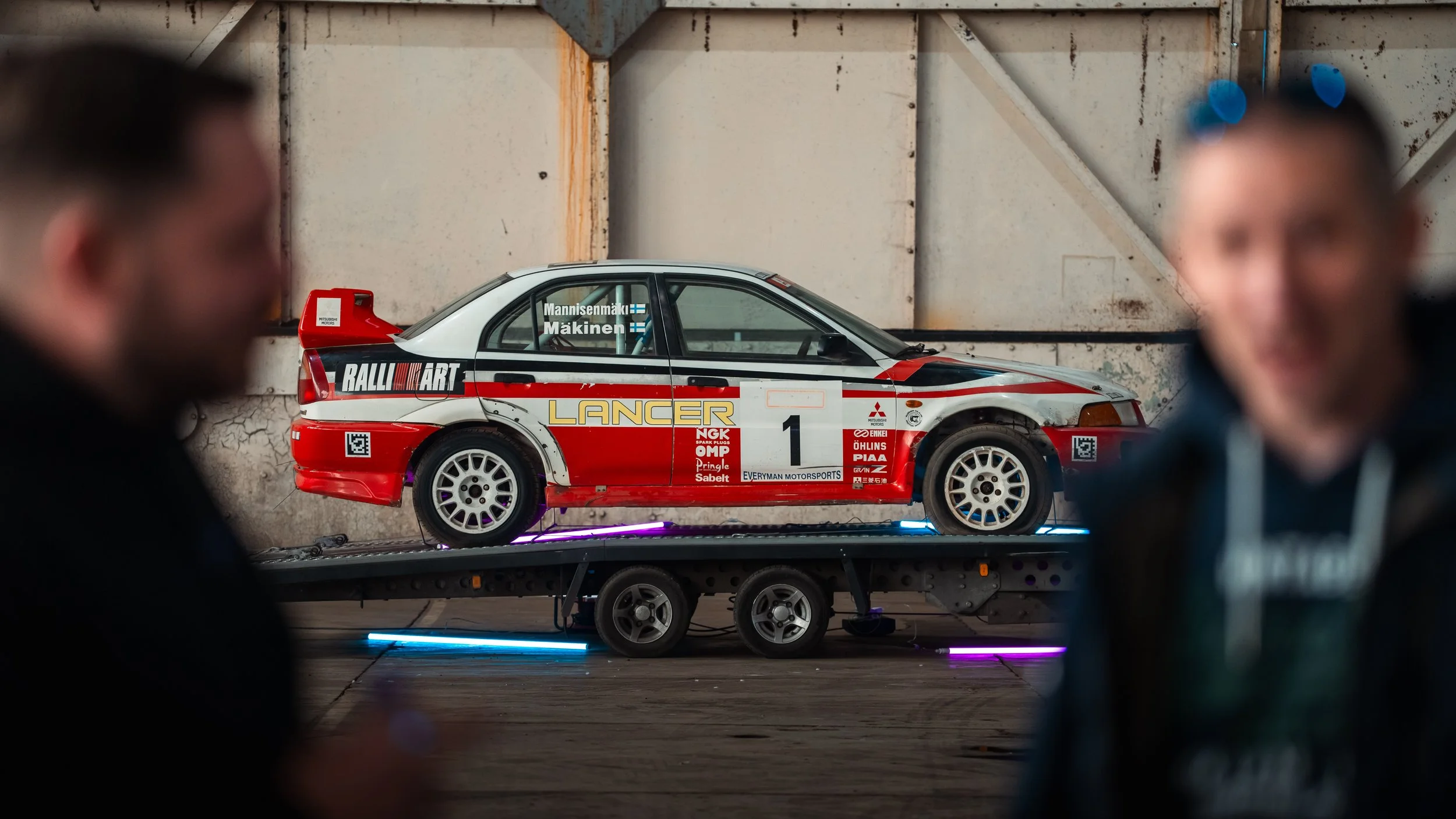 A detailed scale model of a rally car on a dolly, with blurred people in the foreground, in an industrial setting.