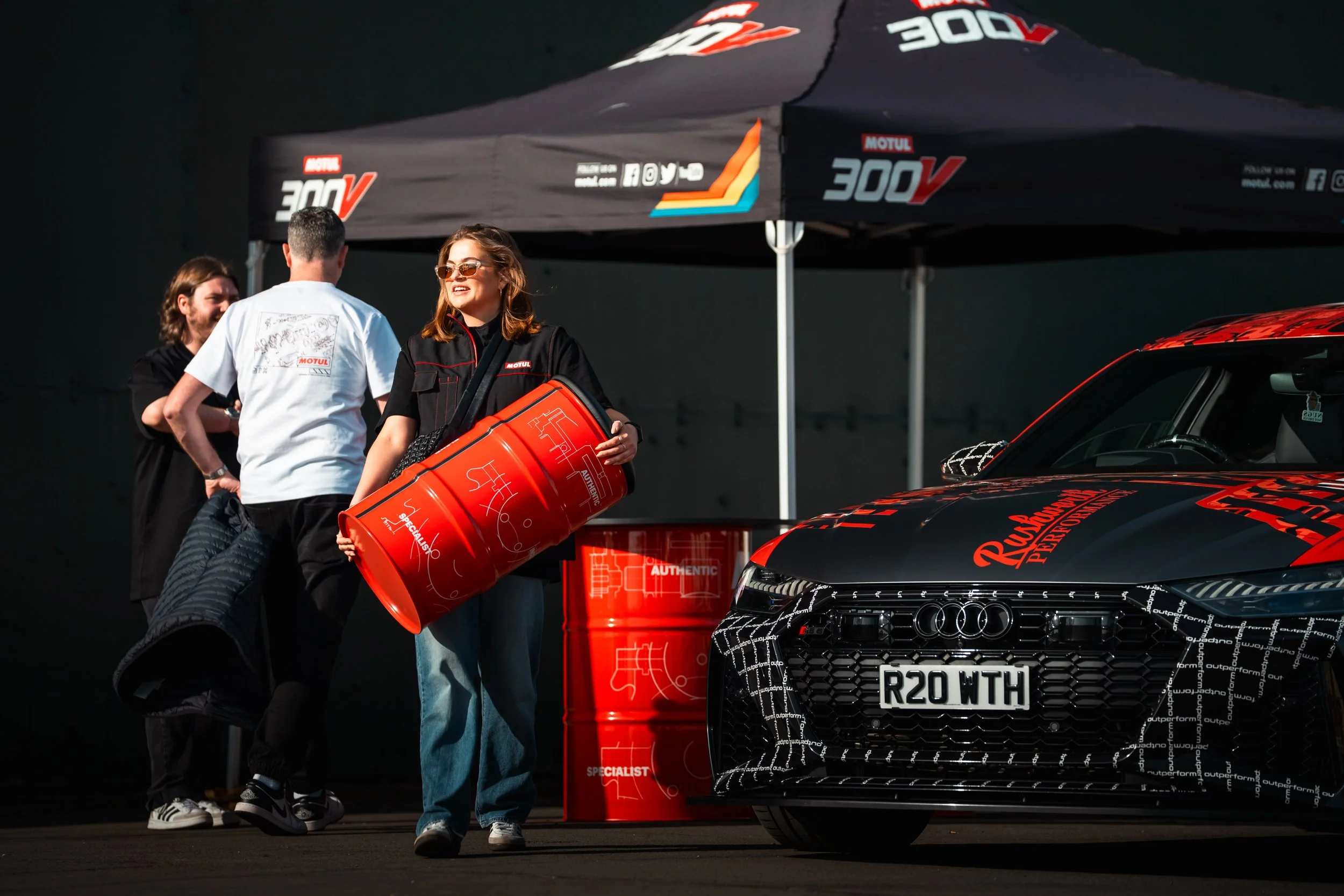 Three people at a motorsports event standing next to a black and red Audi car with aftermarket modifications and a custom license plate, under a black canopy with racing branding.