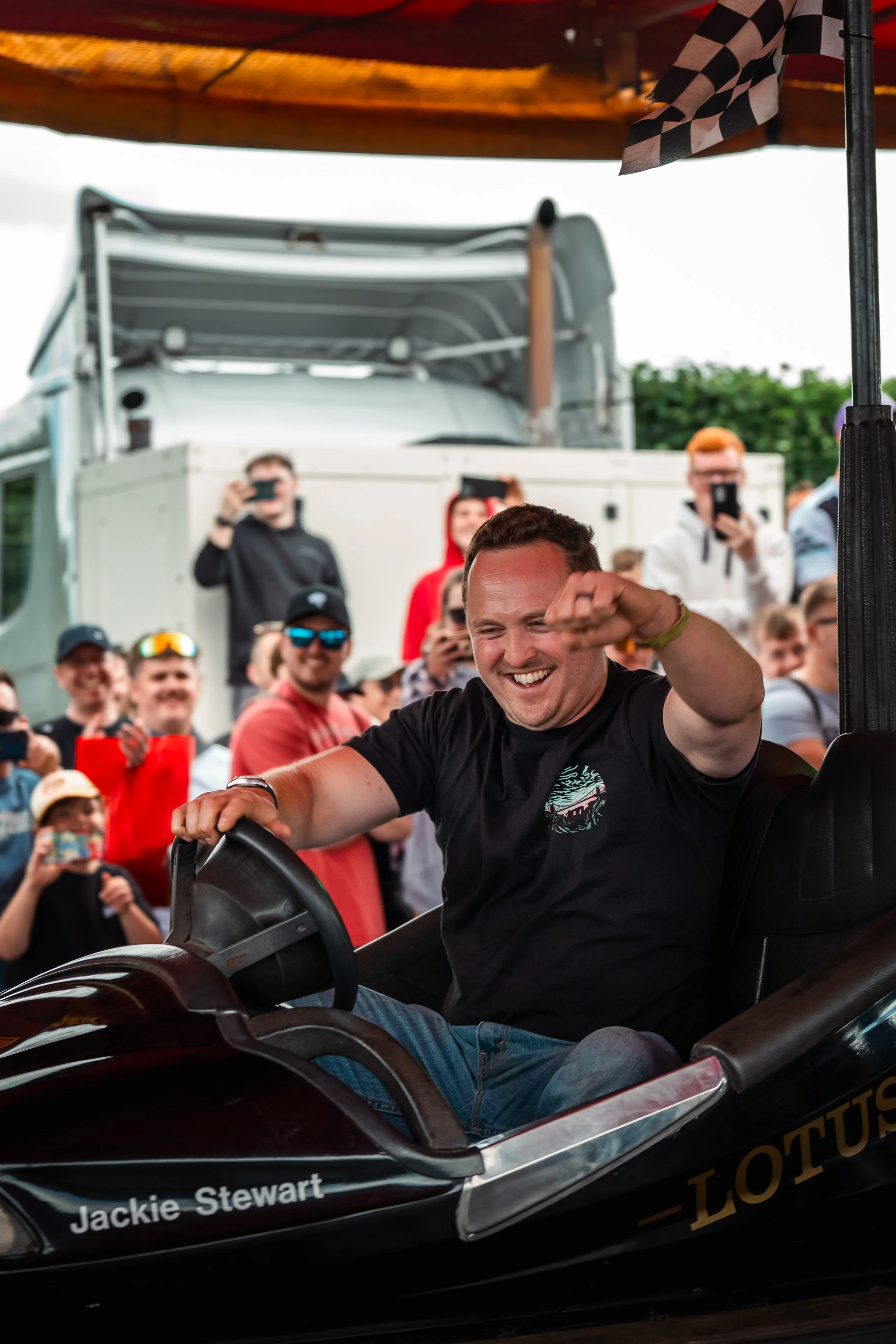 A person with short hair smiling and driving a race car with the name Jackie Stewart on the side at an outdoor event, surrounded by a crowd of spectators taking photos and videos.