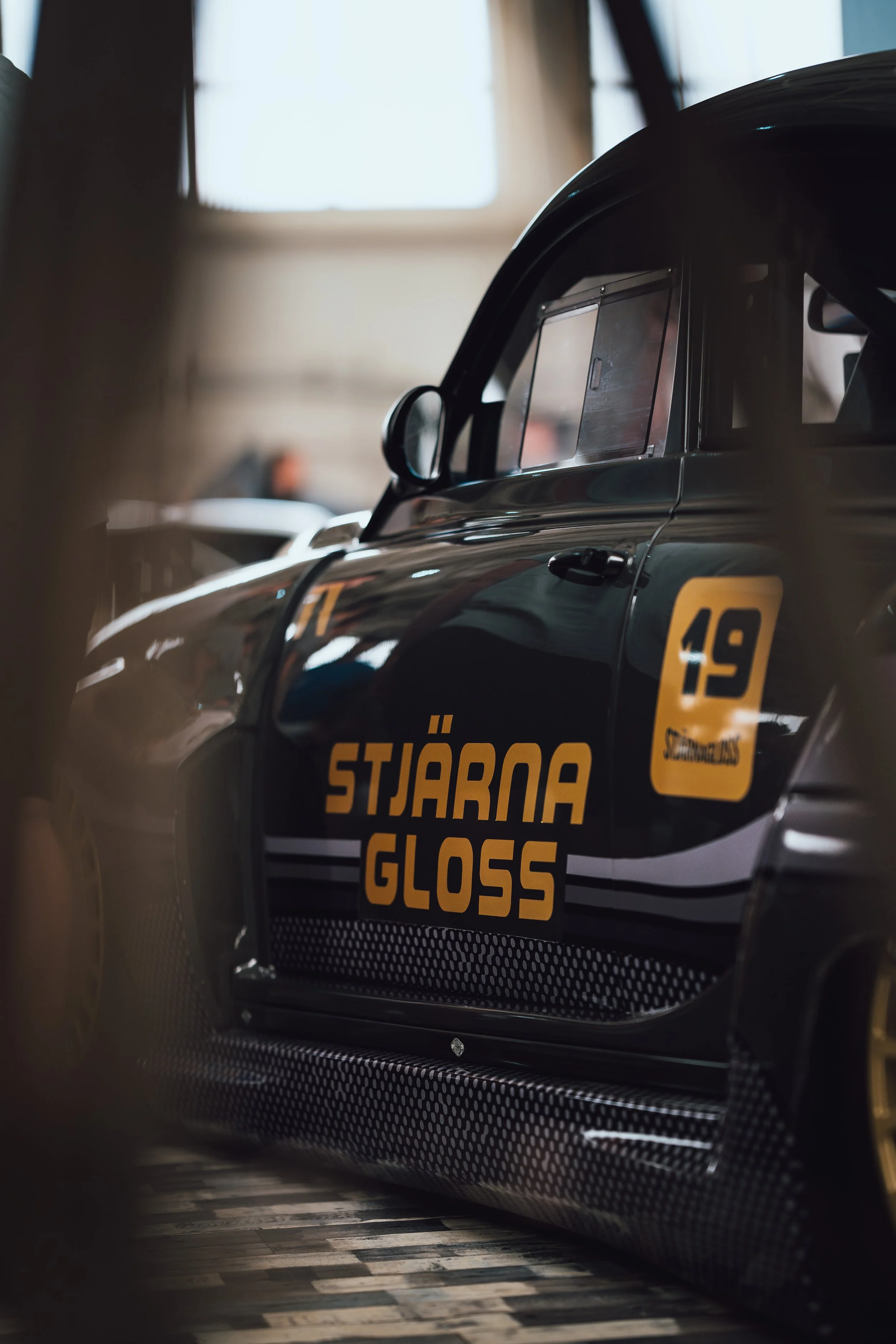 A close-up of a black racing car with yellow and orange graphics, displaying the words "Stjärna GLOSS" on the side.
