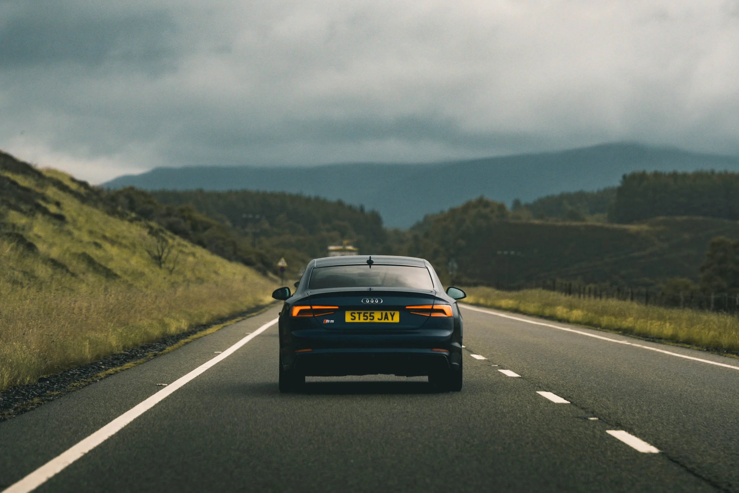 A black Audi S5 driving on an open road through a hilly countryside with trees and shrubs, under a cloudy sky.