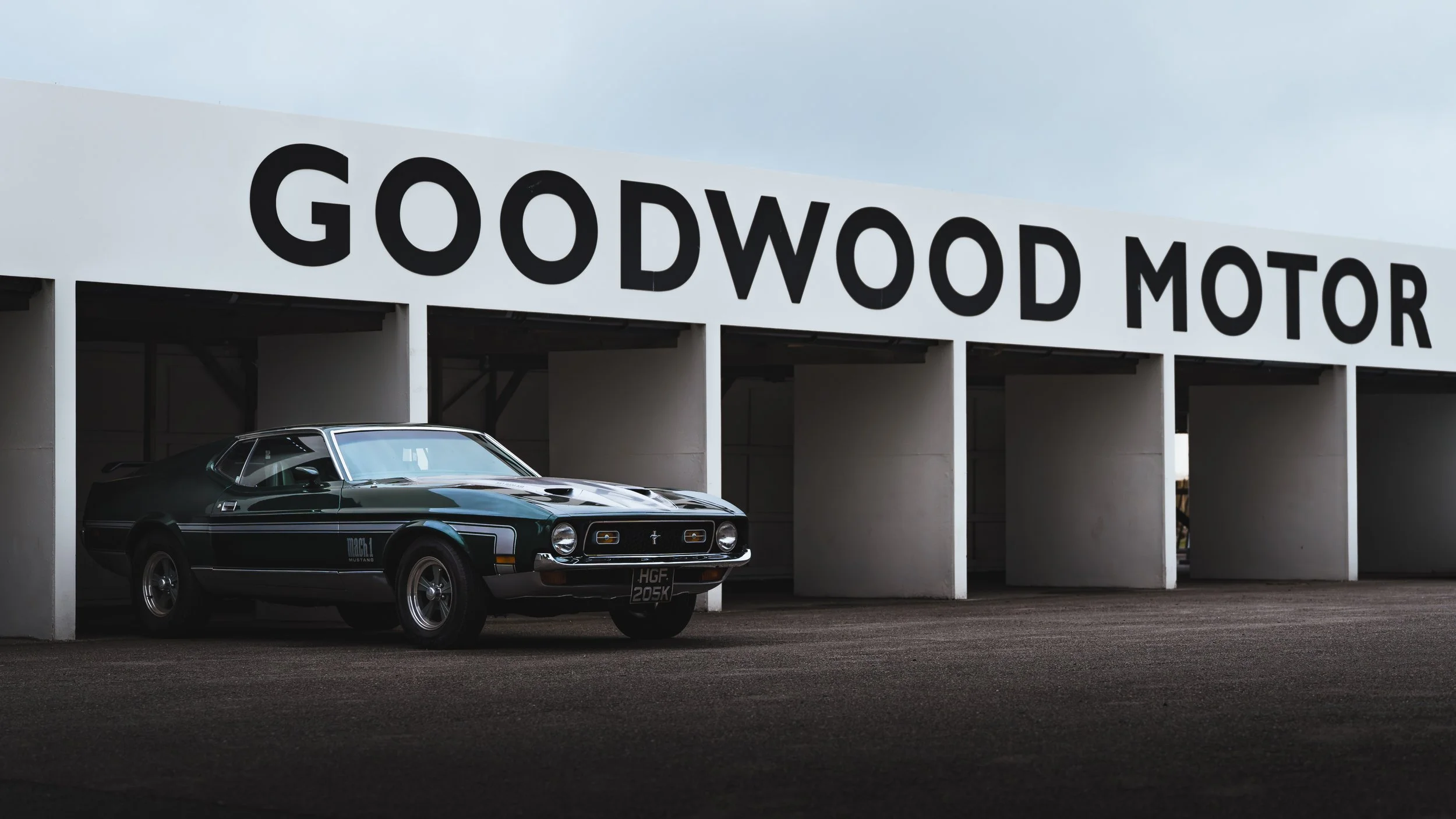 A vintage black Ford Mustang car parked in front of a modern dealership with large black lettering that reads 'Goodwood Motor'.