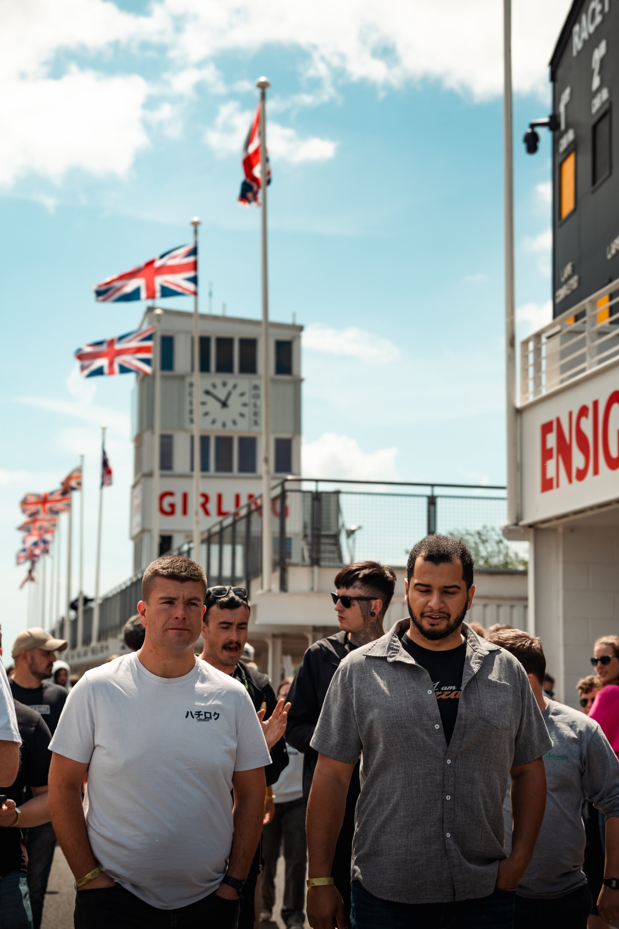 A group of people walking outdoors near a building with flags, a clock tower, and a sign that reads 'GIRL' in the background.