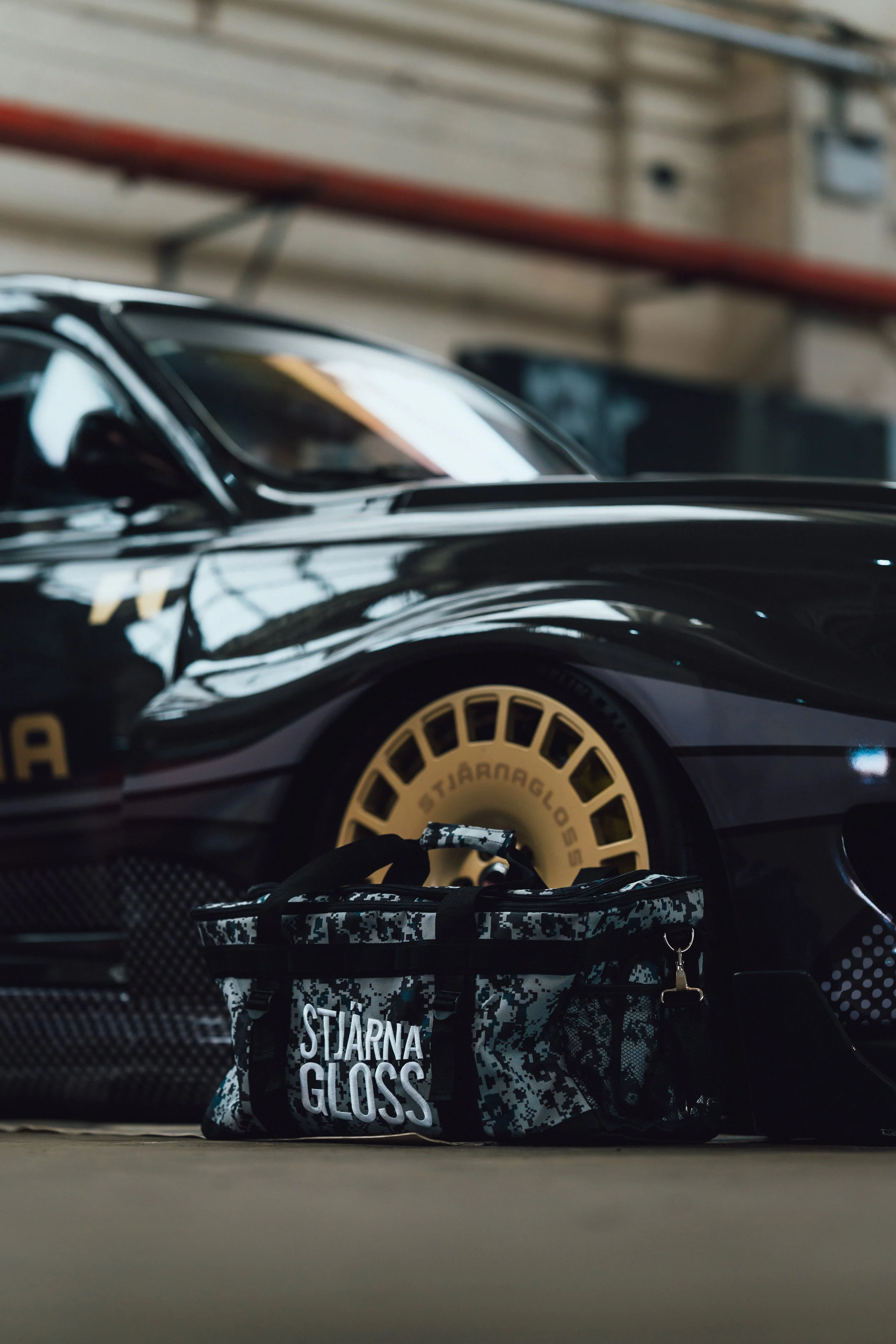 A black racing bicycle with gold wheels parked indoors, with a black and white patterned bag with the words 'Starna Gloss' placed in front of it on the floor.