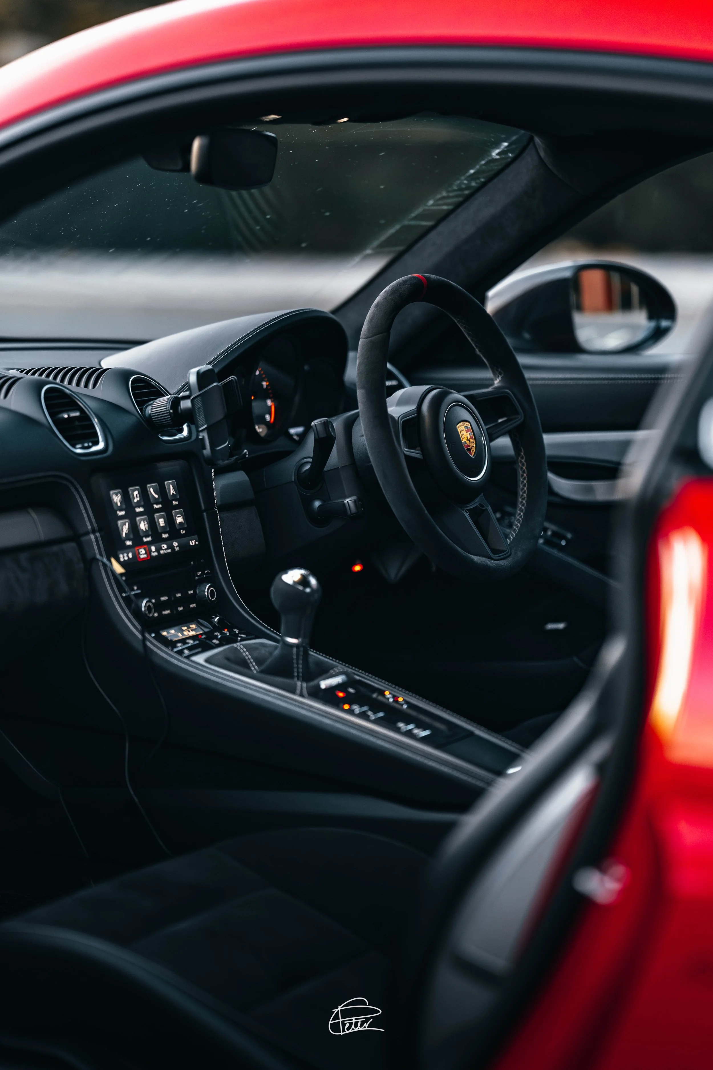 Interior of a red Porsche car showing the dashboard, steering wheel with Porsche logo, gear shift, dashboard screen, and side mirror, captured through the car's window.