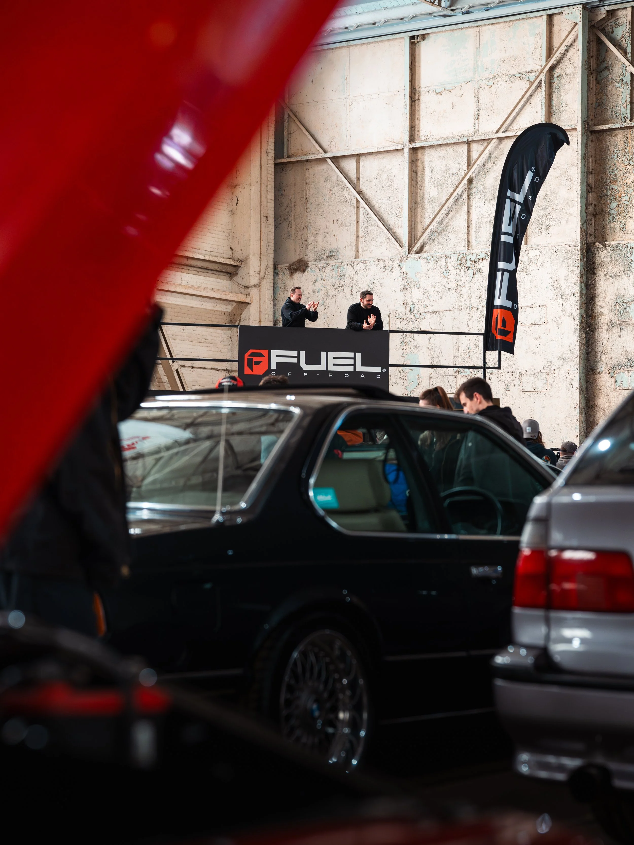 Two men standing behind a black Fuel Off-Road banner, clapping and congratulating at an automotive event inside an industrial building, with several cars parked in the foreground.