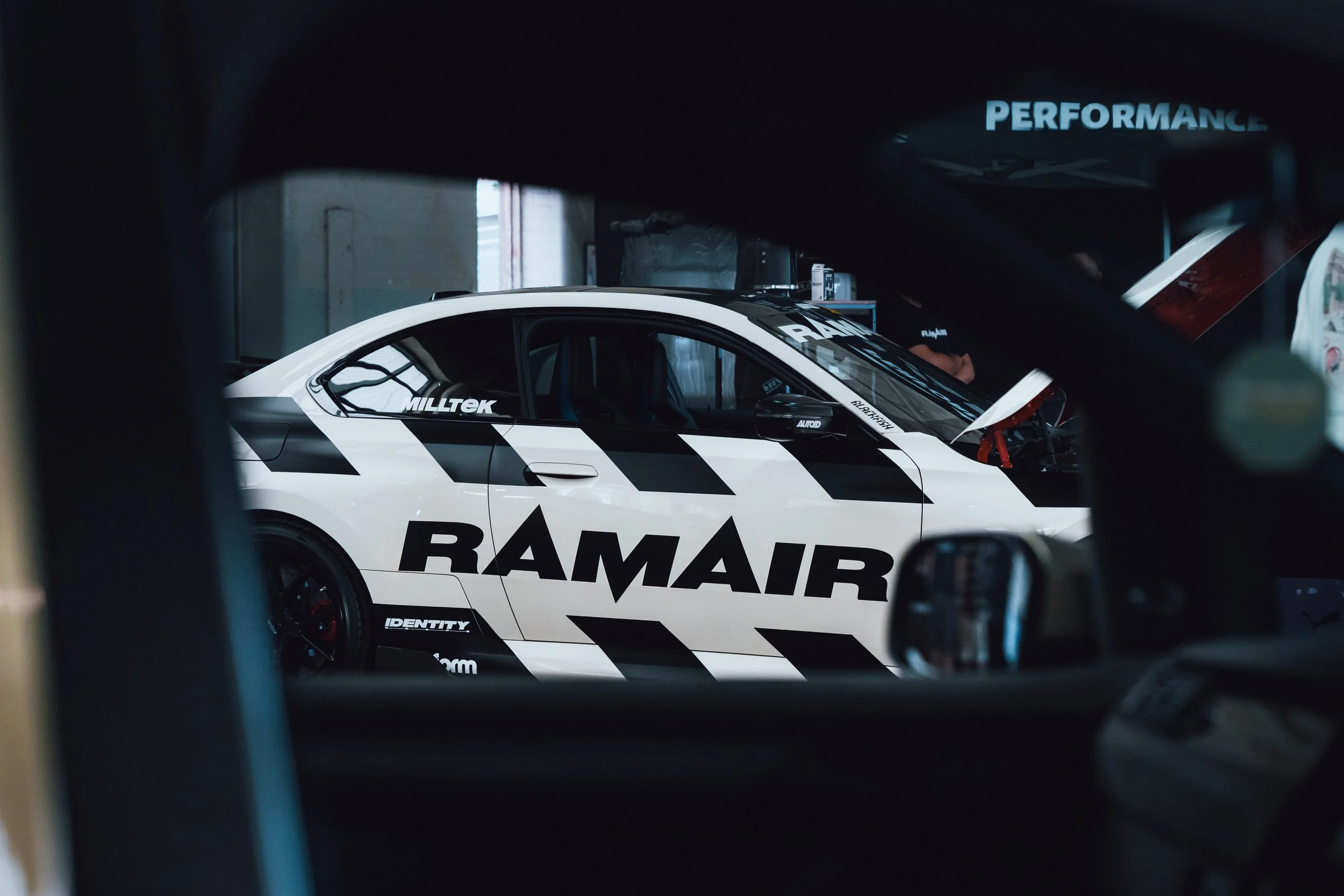 A race car inside a garage, with the word 'RAMAIR' on its side and hood, and the brand logo 'MILLTEK' near the front. Someone is working on the car in the background, and a blue sign with the word 'PERFORMANCE' is visible.