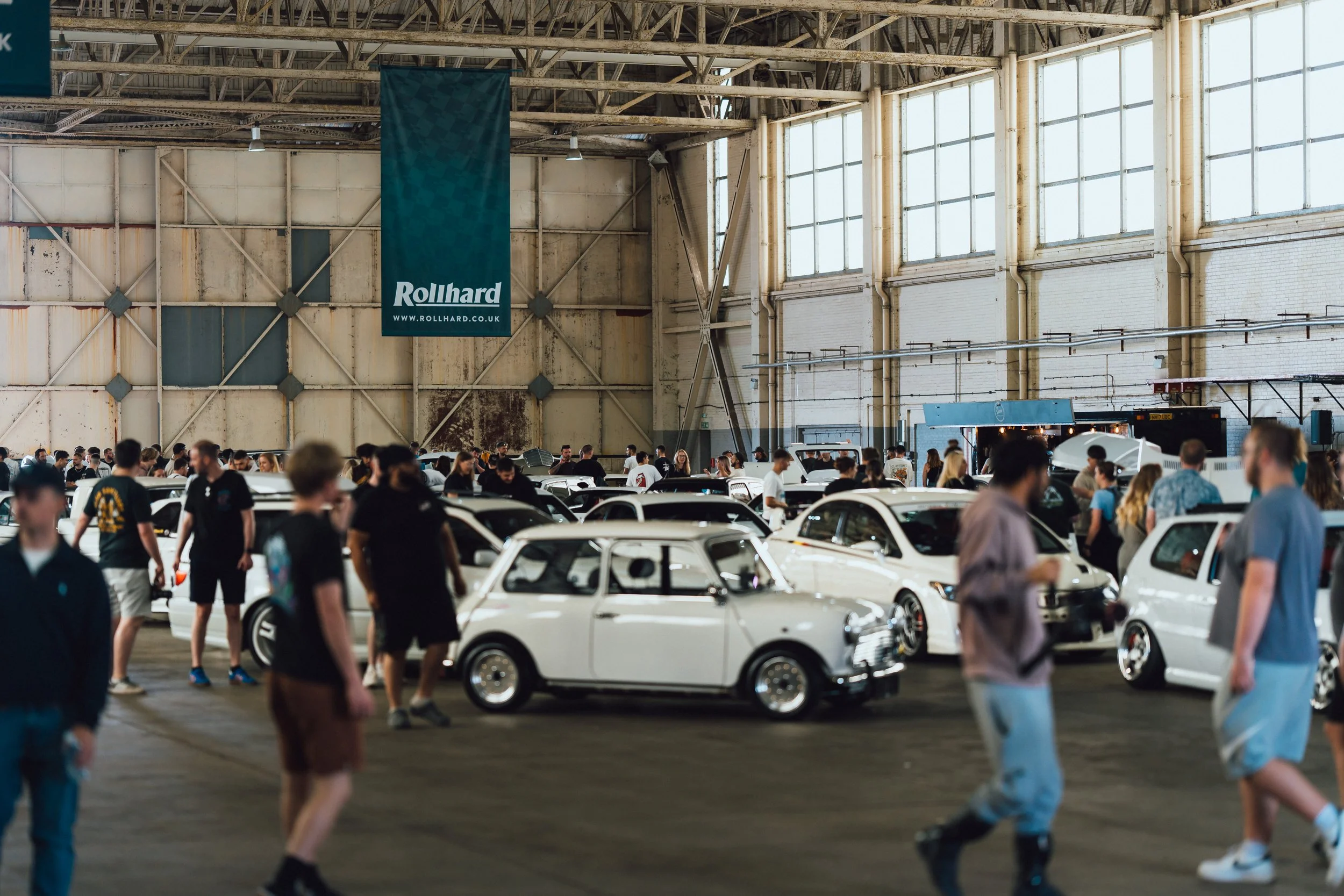 An indoor car exhibition with numerous classic cars and many visitors walking around. There is a large banner hanging from the ceiling with the website 'rollhard.co.uk' and the name 'Rollhard' on it.