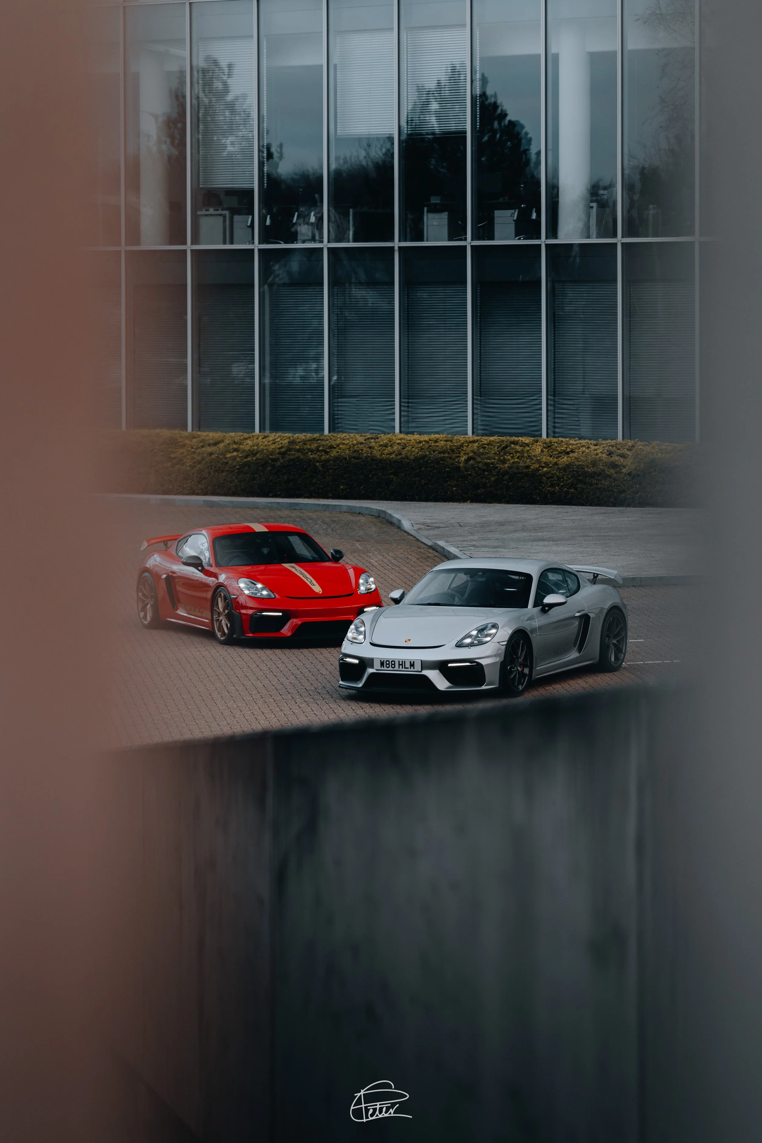 Two luxury sports cars, a red one and a silver one, parked on a paved lot in front of a modern glass building with trees reflected in the windows.