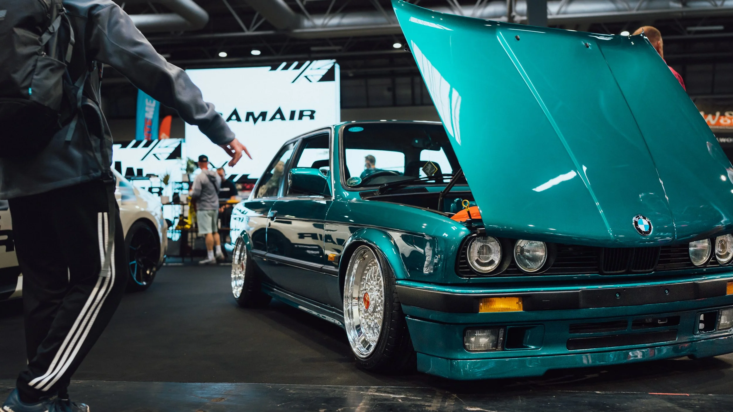 A teal vintage BMW car on display at an automotive exhibition with its hood open, surrounded by people and other vehicles.