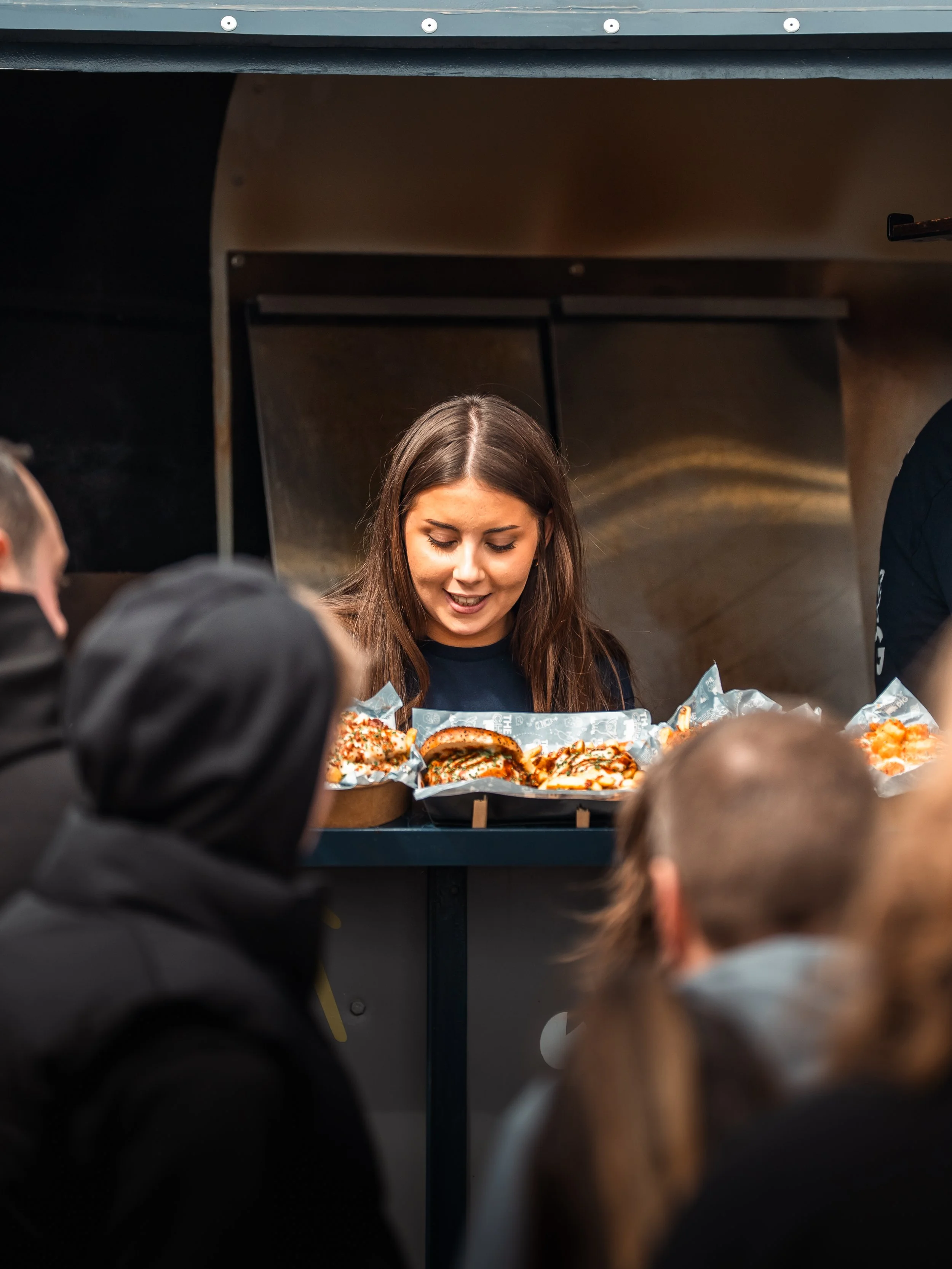 A woman with long brown hair smiling and looking down behind a display of sliced pizza with various toppings, with a dark background and parts of other people visible.