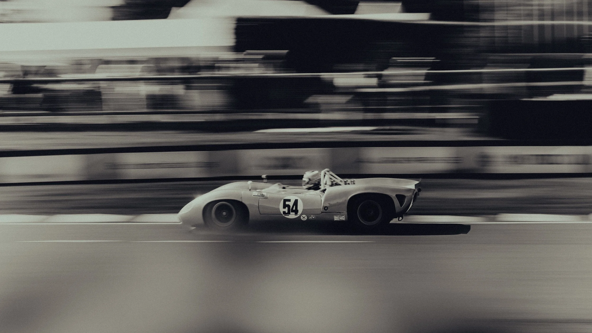 A vintage race car with the number 54 on its side speeds along a racetrack, with motion blur in the background.