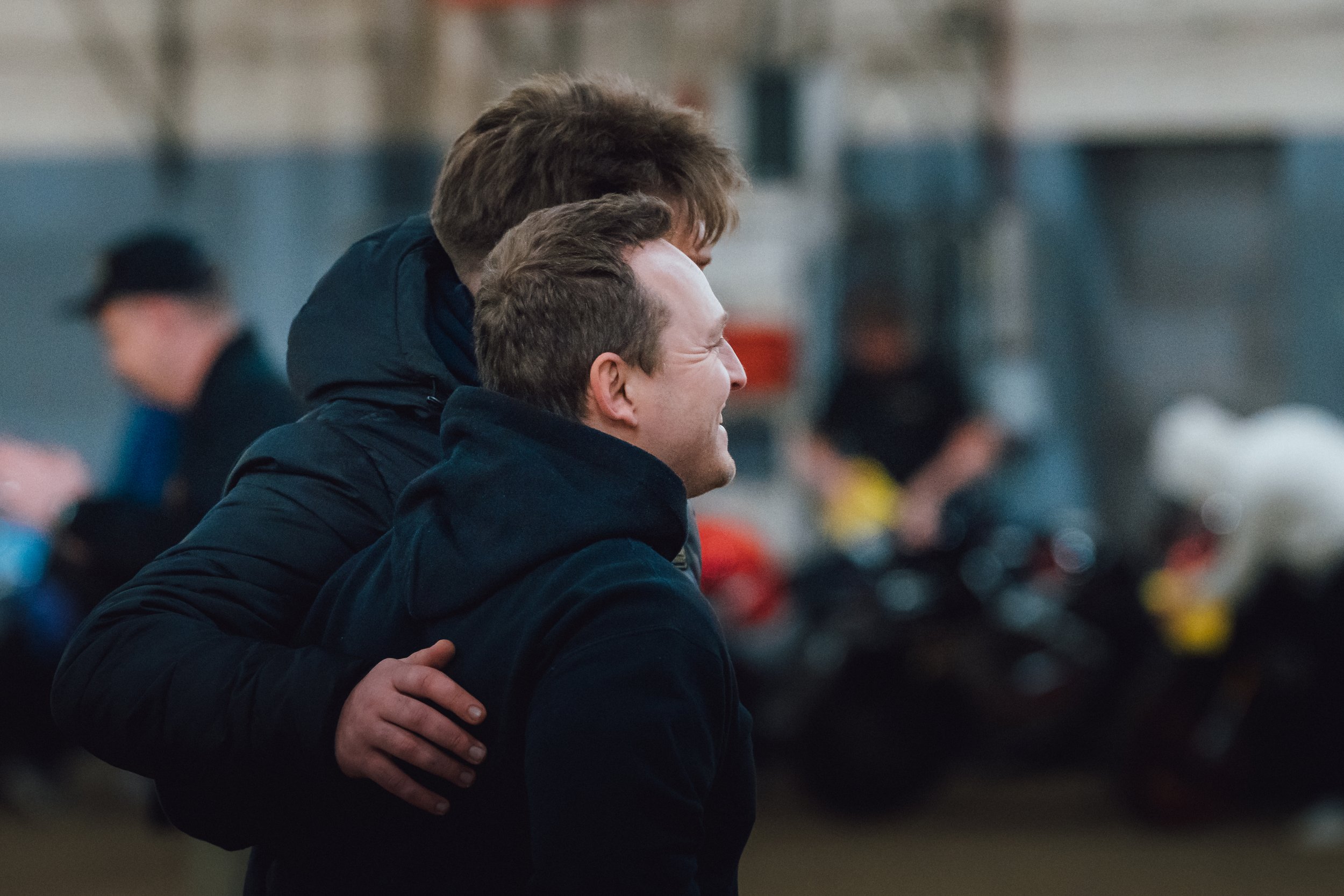 Two young men embracing each other, one with his hand on the other's shoulder, smiling warmly. The background is blurred, suggesting they are outdoors or in a busy environment.