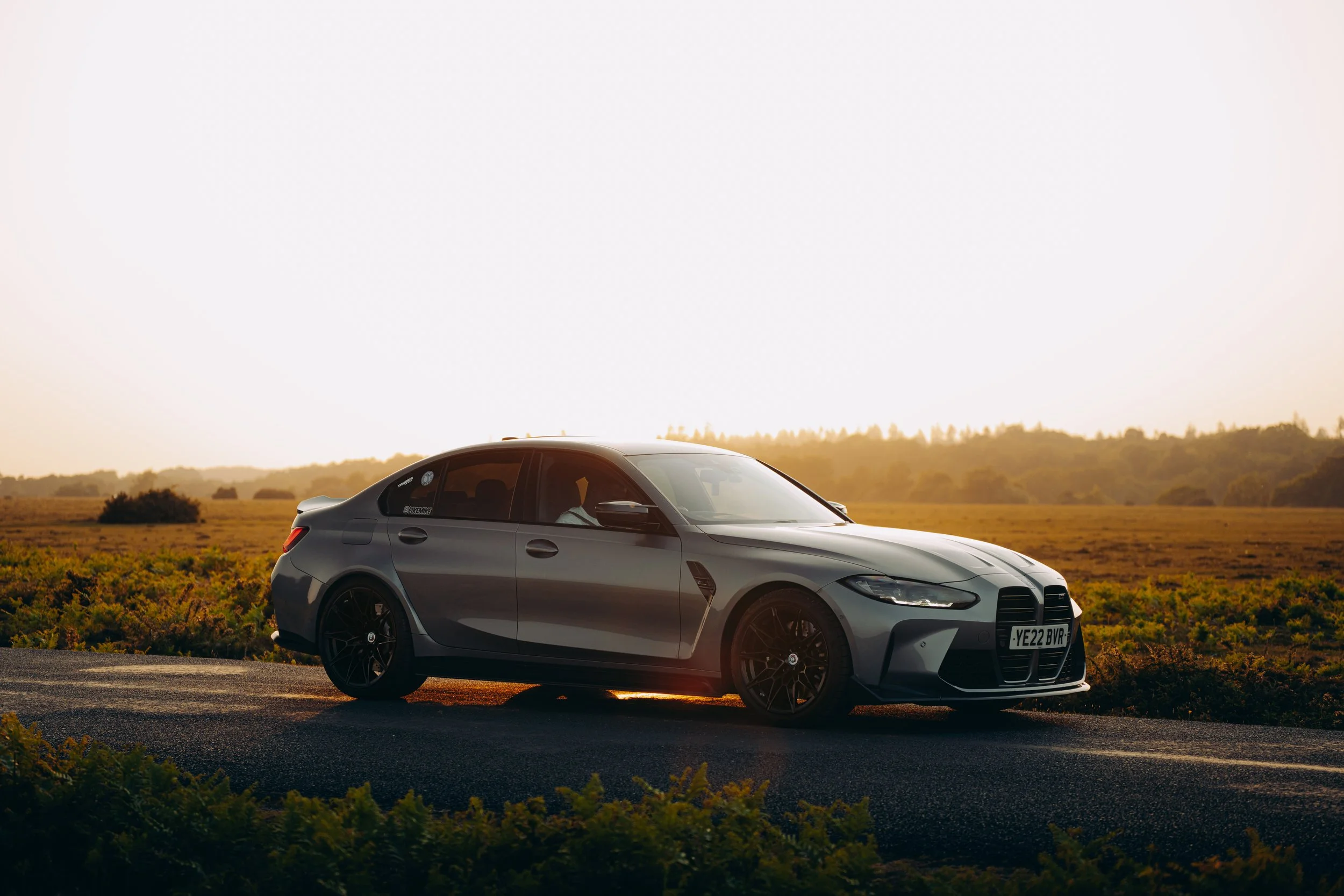 A sleek silver sports car parked on the side of a road, with a backdrop of a picturesque countryside landscape during sunset.