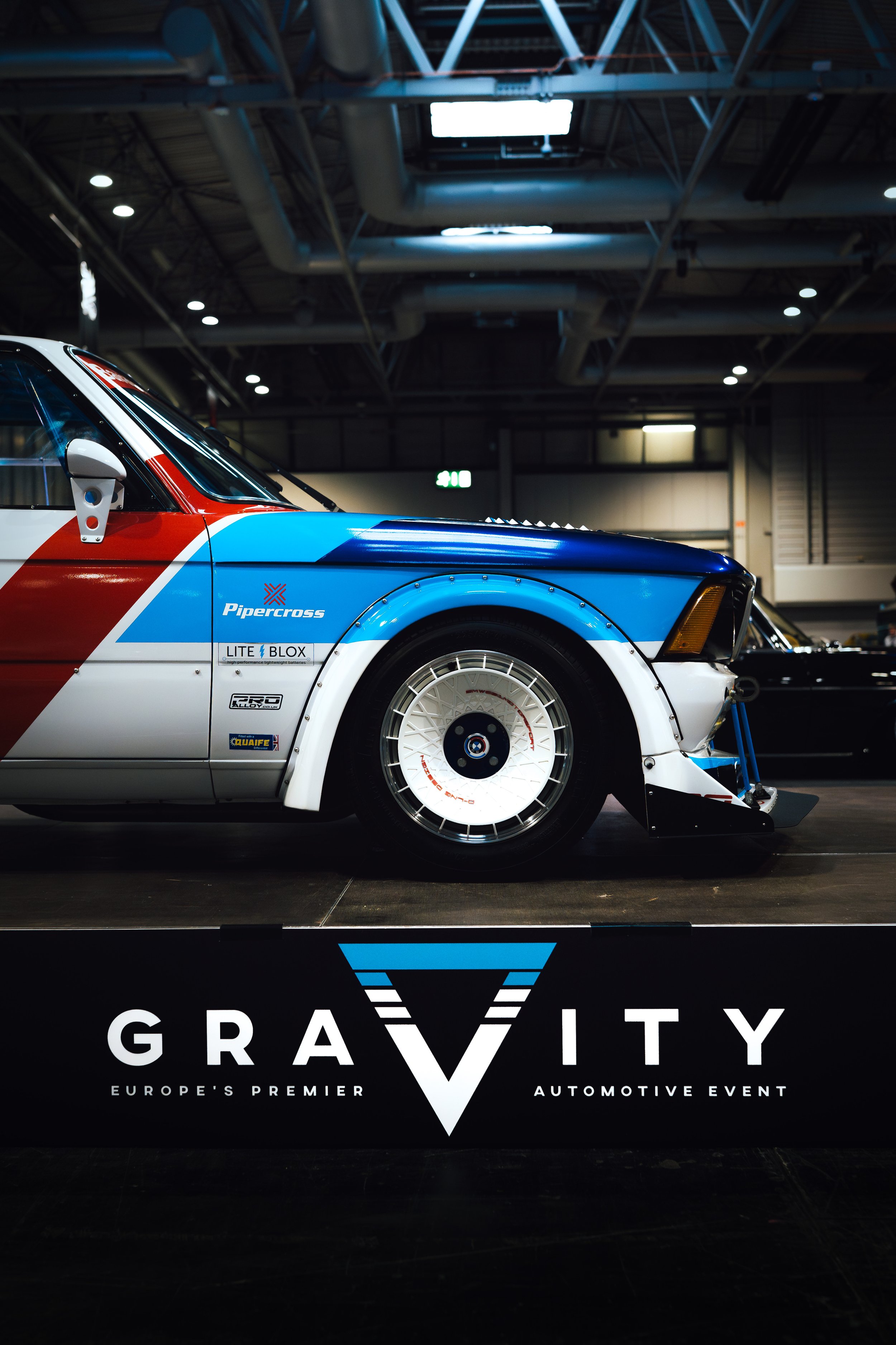 Close-up of a custom race car with patriotic livery in blue, white, and red colors, displayed at an automotive event with a sign reading 'GRAVITY' and the subtitle 'Europe's Premier Automotive Event'.