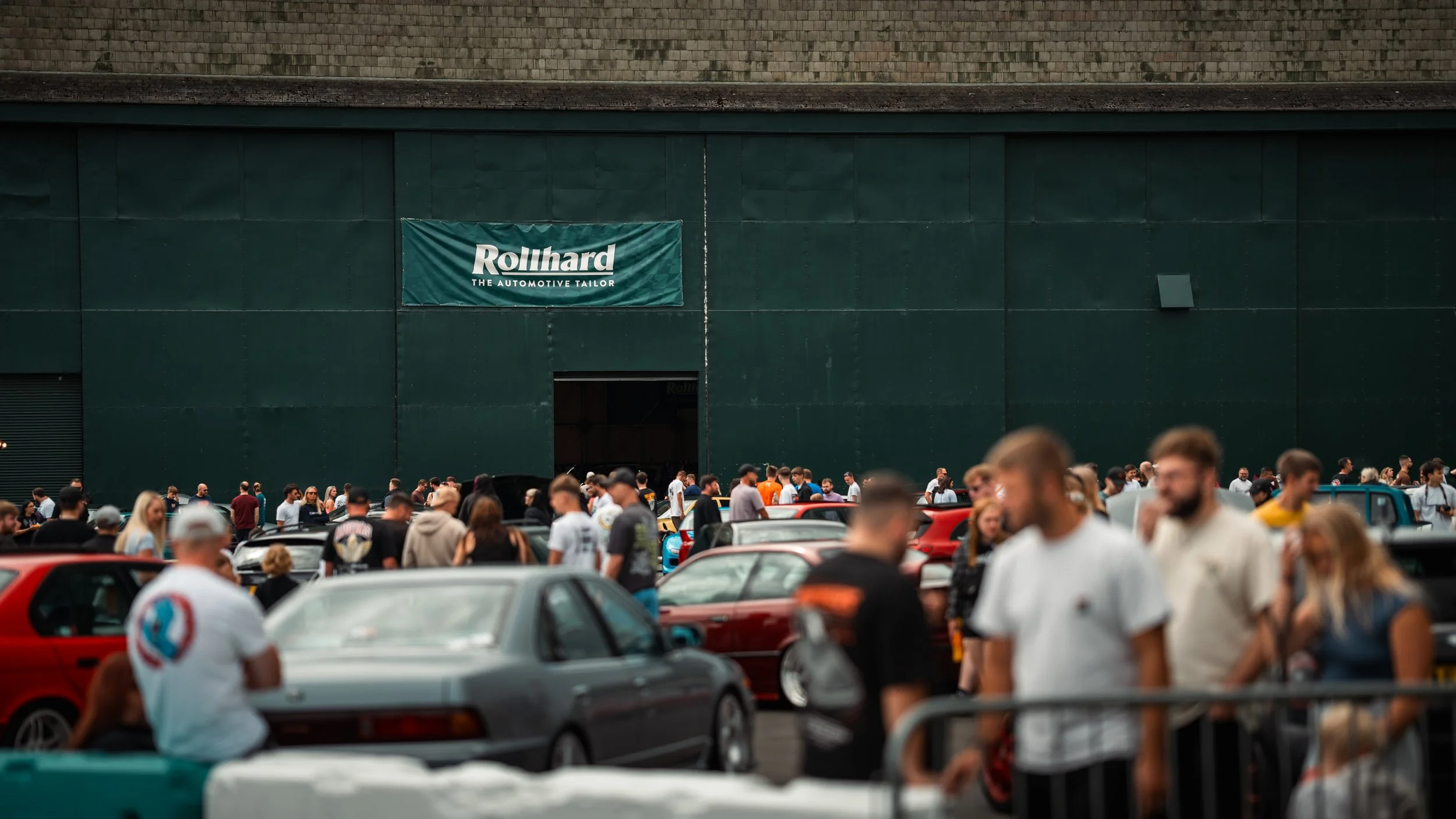 A crowd of people gathered at a car event outside a building with a large green sign reading "Rollhard: The Automotive Tailor".