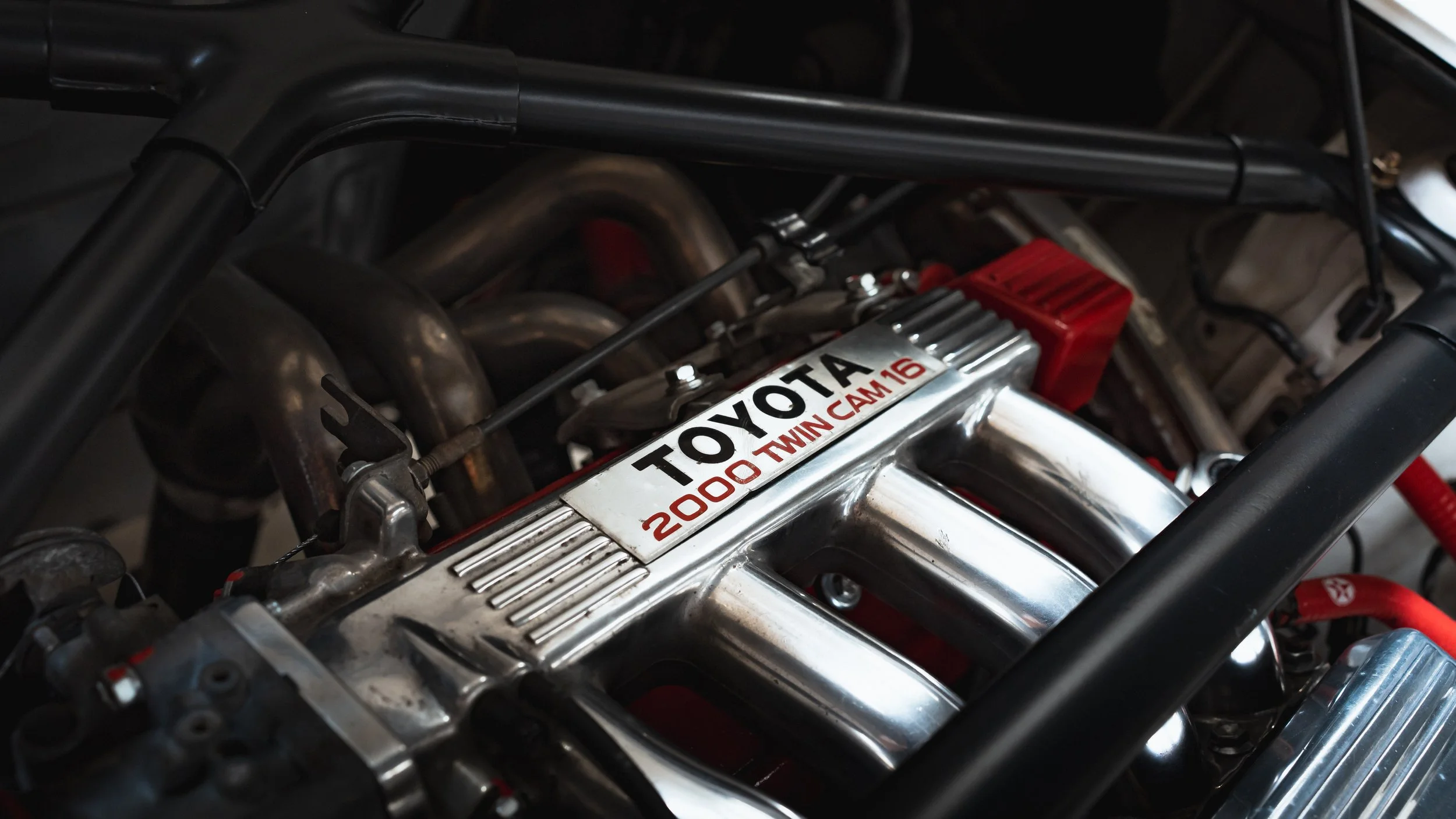 A close-up of a race car engine with a Toyota 2000 twin cam 16 sticker on the valve cover and various metal pipes and parts surrounding it.