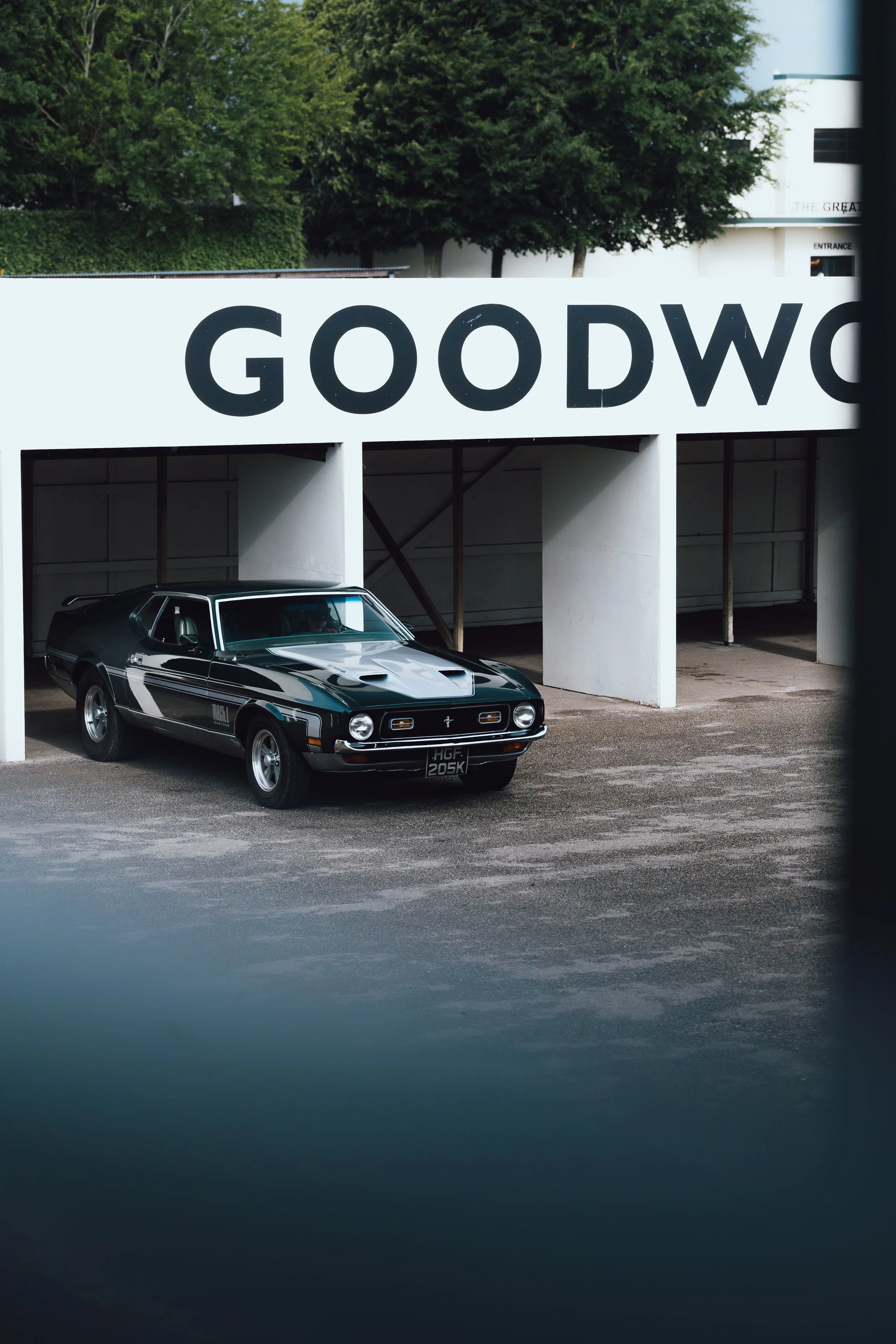 A black vintage car parked under a white building with large black lettering spelling "GOODWOOD" and with green trees in the background.