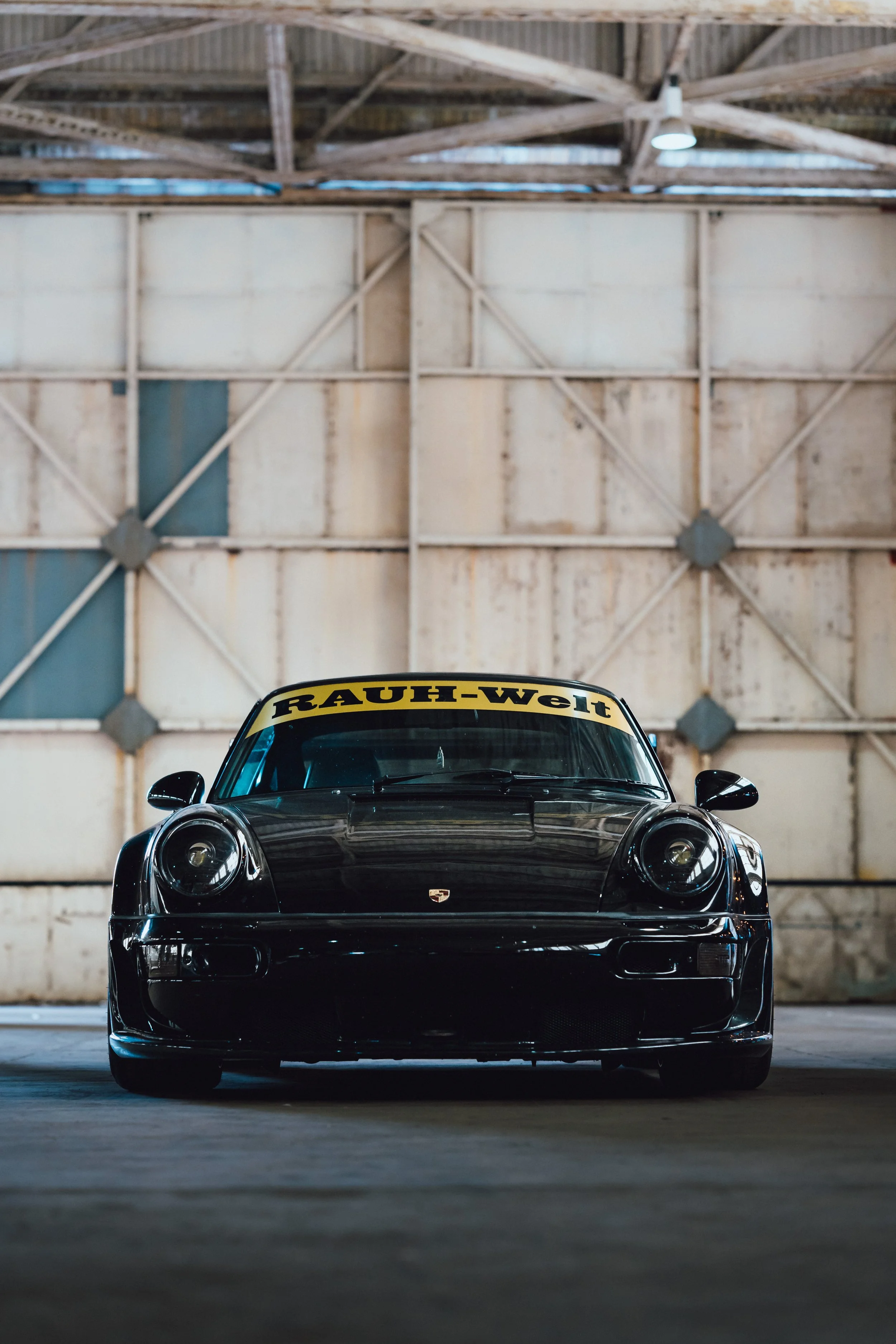Front view of a black Porsche sports car with a RAUH-Welt sticker on the windshield, parked inside a large industrial warehouse with exposed wooden beams and a high ceiling.