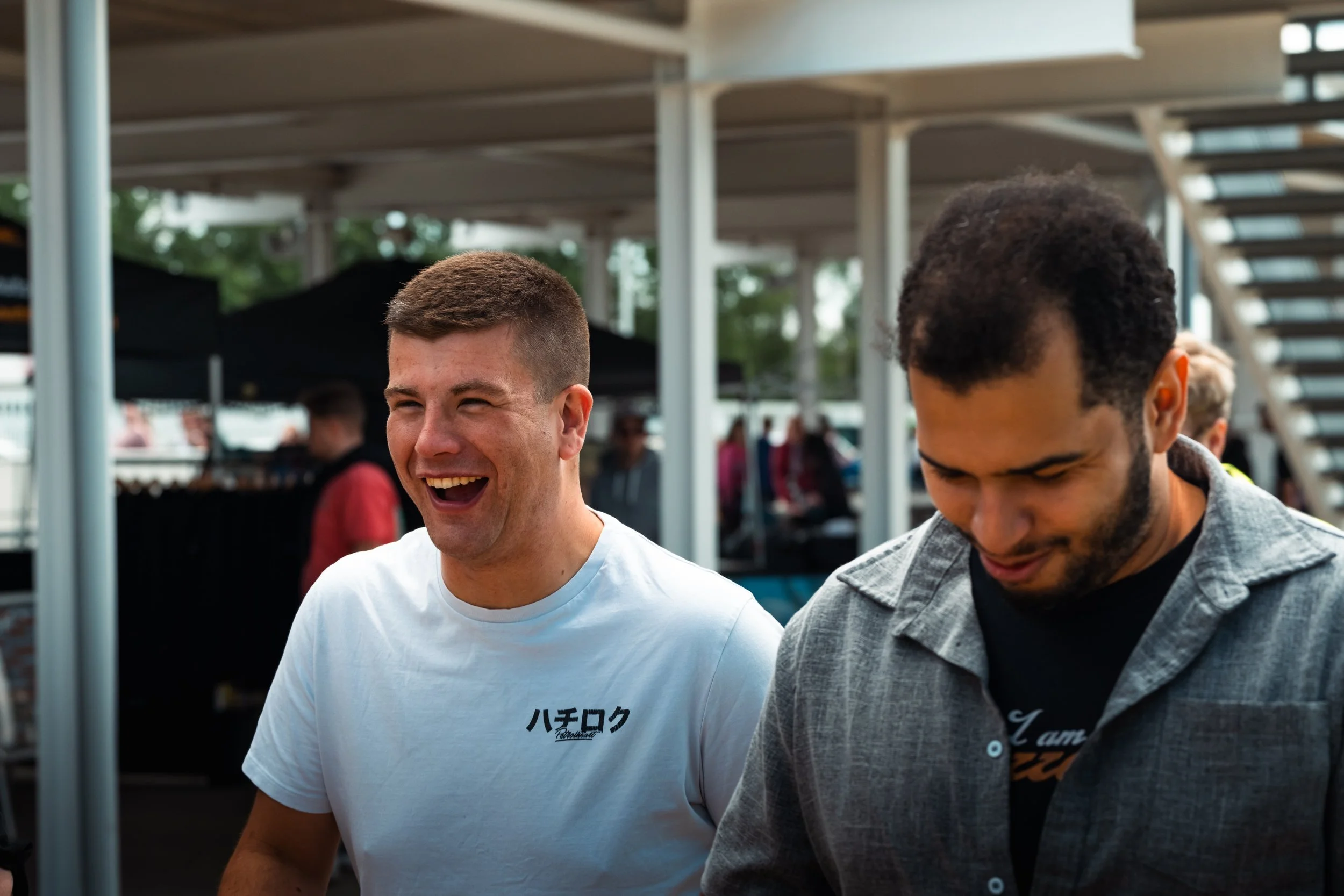 Two young men standing outdoors, smiling and looking down, with people and a staircase in the background.