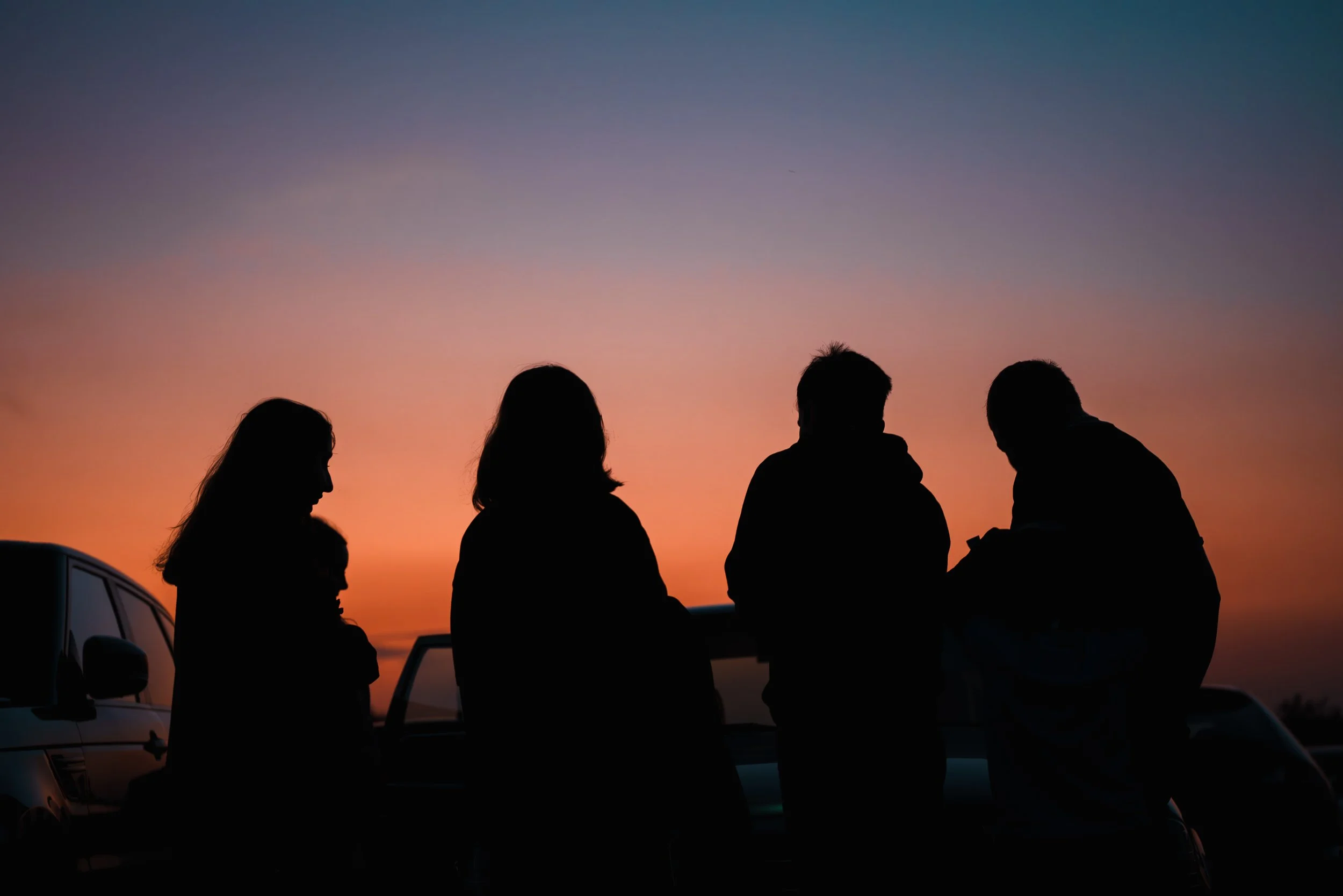 Silhouettes of five people standing near a car during sunset, with a colorful sky in the background.