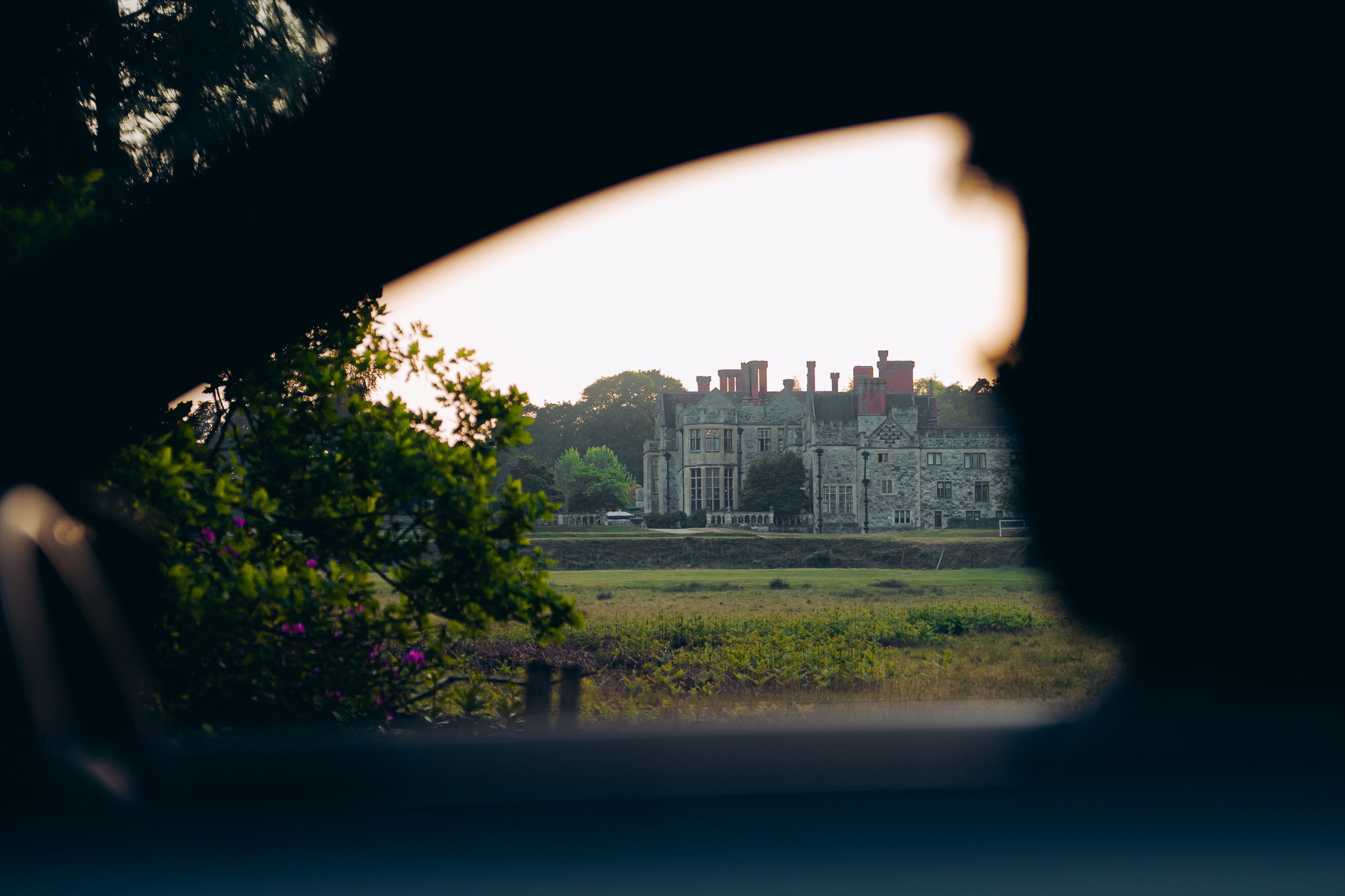 View of a grand, historic mansion with multiple chimneys, seen through a small opening framed by foliage and dark tree branches at sunset.