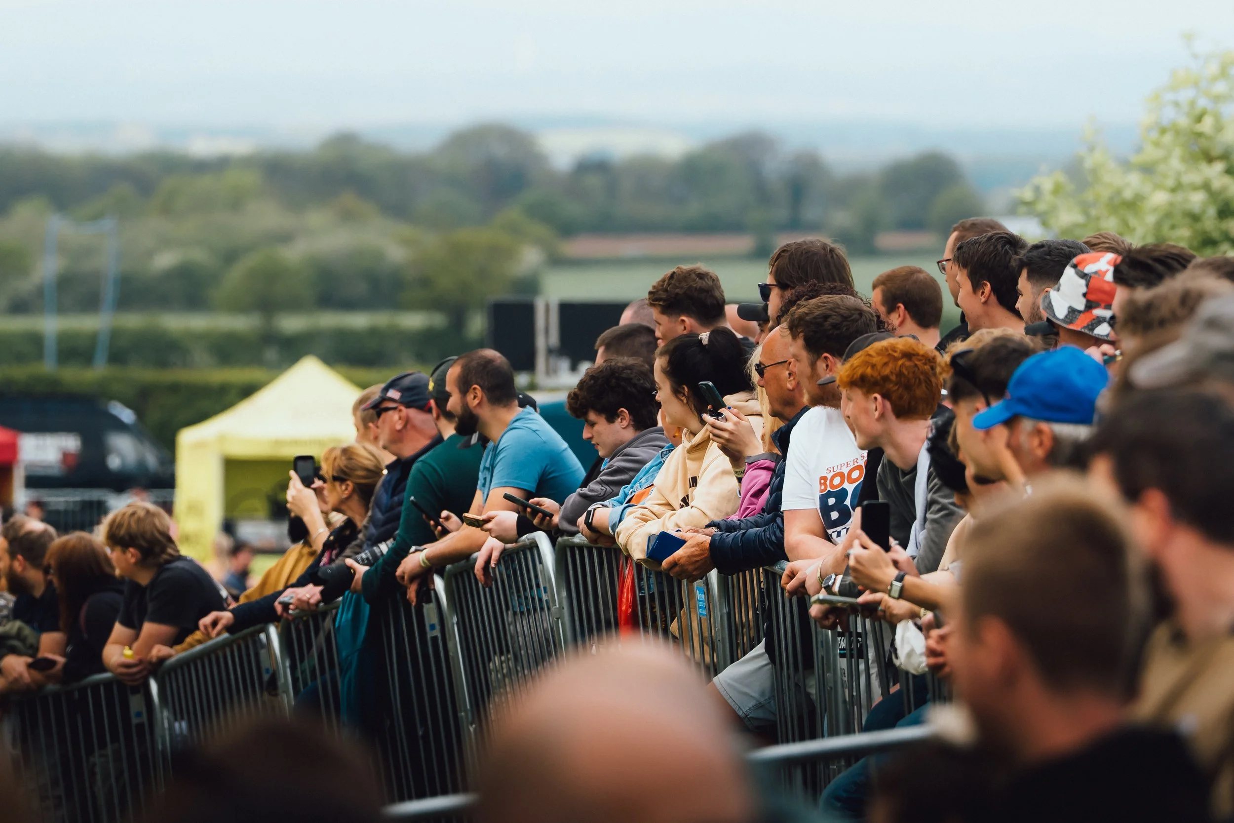 Crowd of people sitting behind metal barricades at an outdoor event, some using smartphones, with green trees and a cloudy sky in the background.
