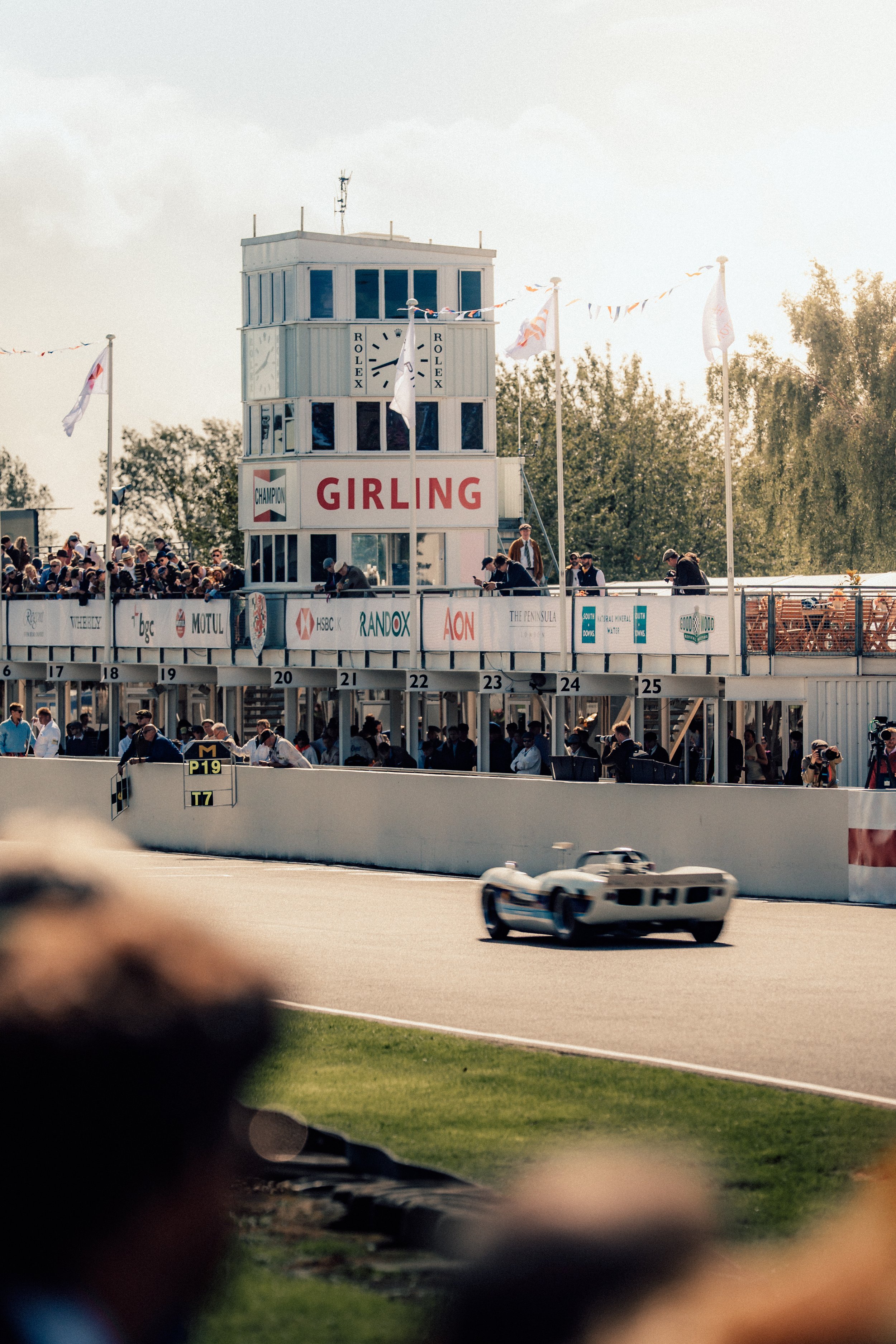 A vintage race car speeding on a race track, passing a crowd of spectators and a control tower with the Rolex logo.