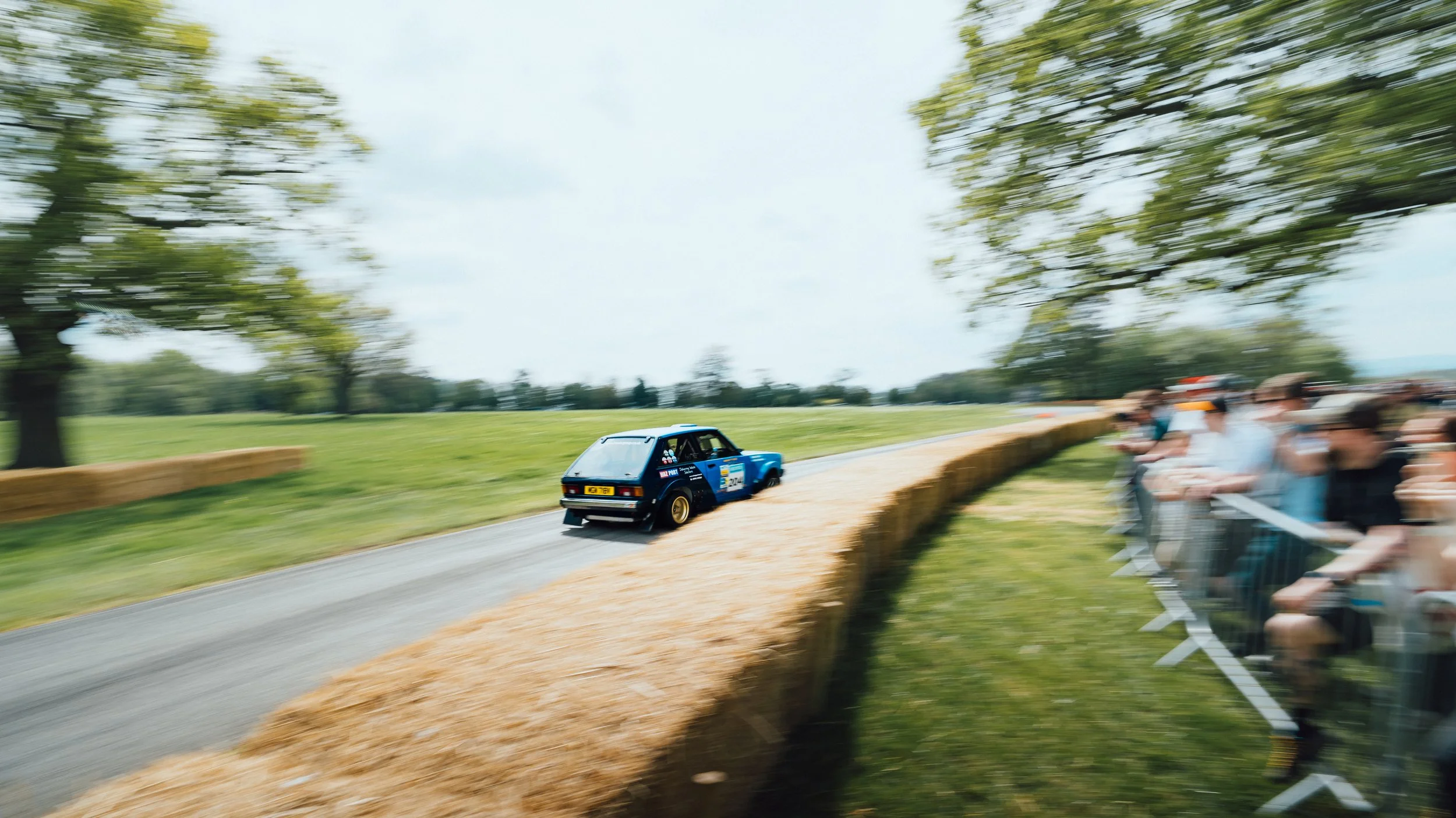 A blue rally car racing on a dirt track with spectators on the side, trees in the background, and motion blur indicating speed.