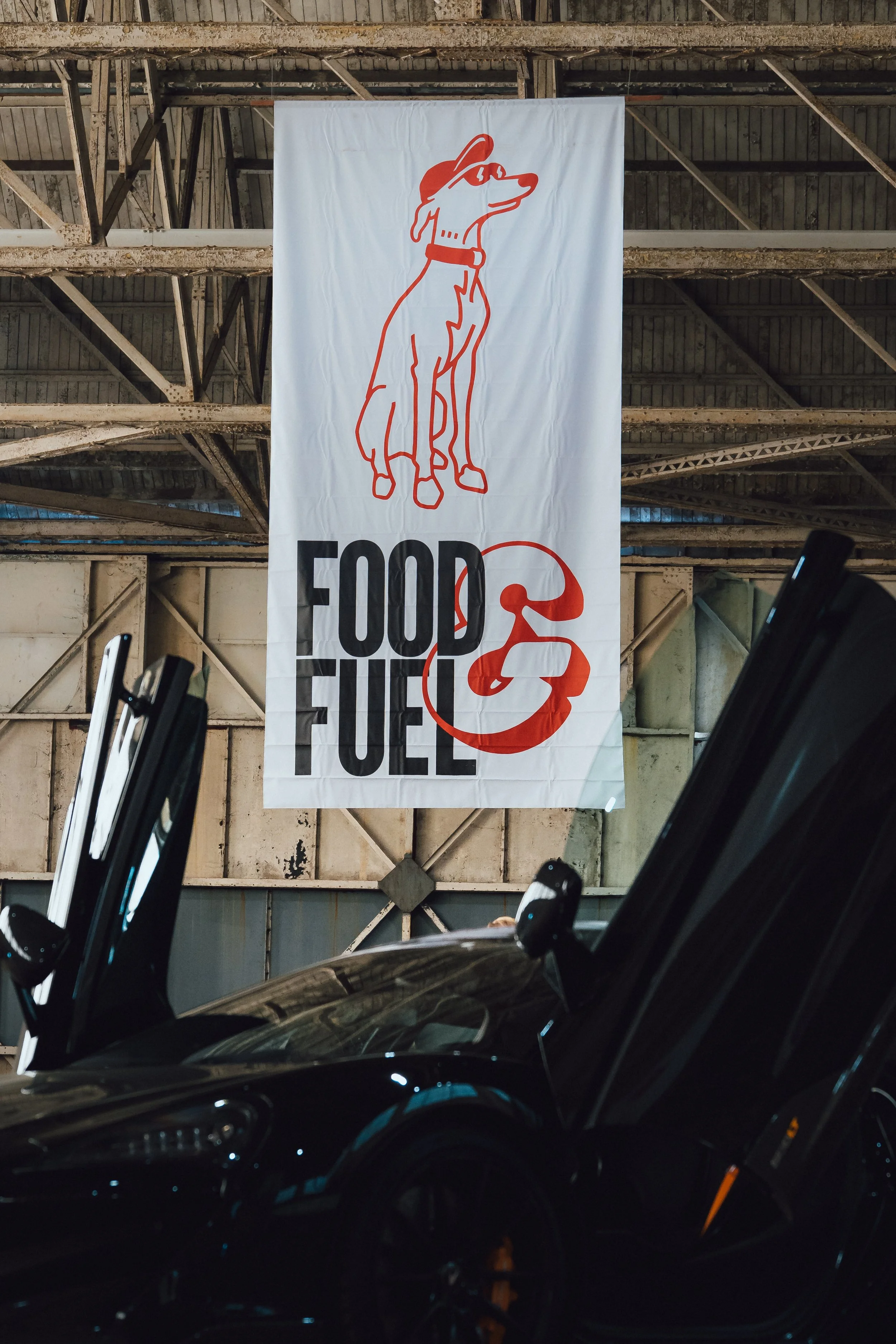 Large banner hanging in an industrial warehouse or parking structure with a line drawing of a dog wearing a baseball cap and glasses, and the text 'FOOD FUELED' with a red circular design around the text.