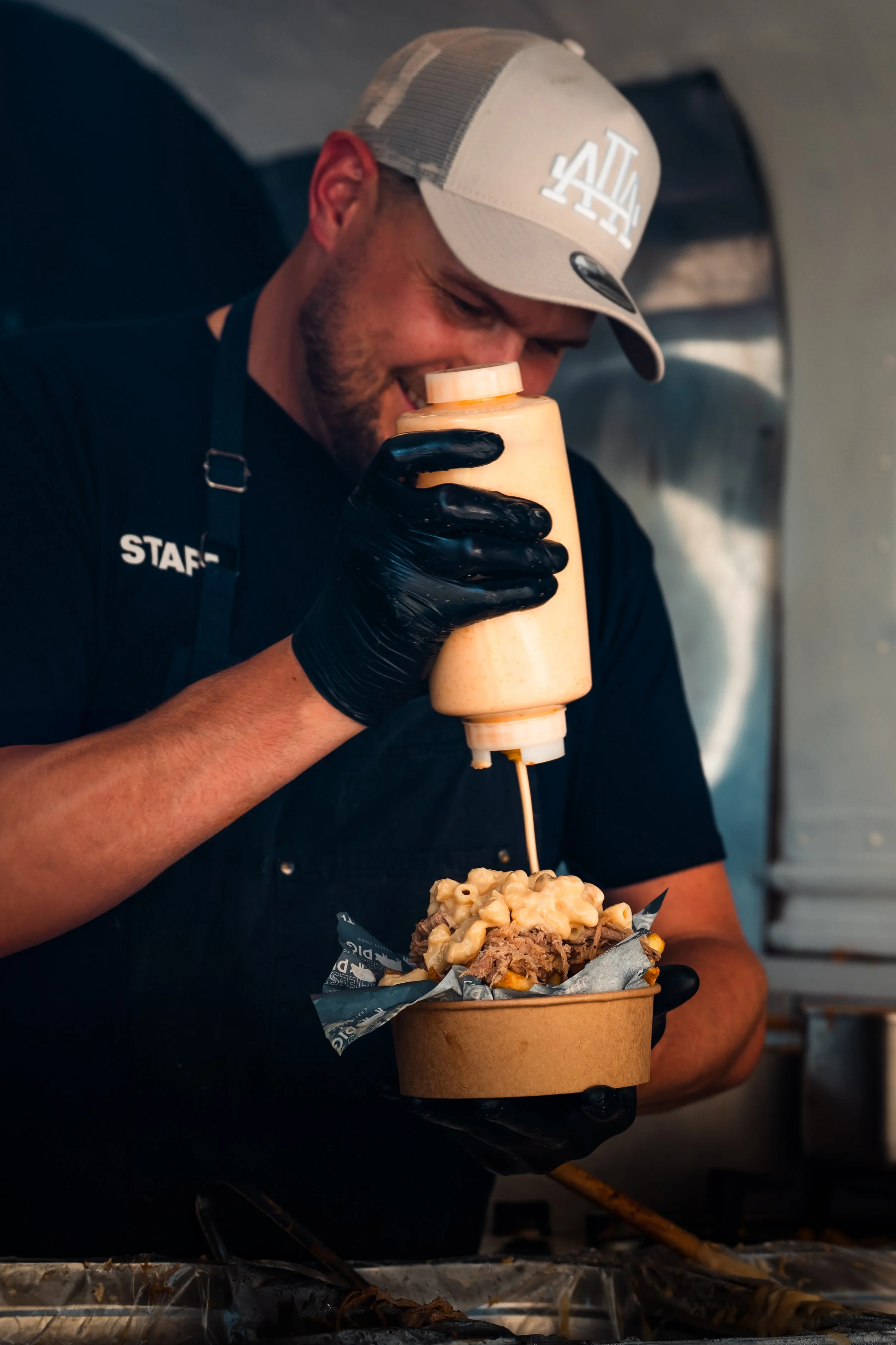 A man wearing a beige Los Angeles baseball cap, black gloves, and a black shirt labeled 'STAFF' is smiling while pouring sauce onto a large food item in a brown paper container. The food item includes macaroni and cheese, shredded meat, and other ing