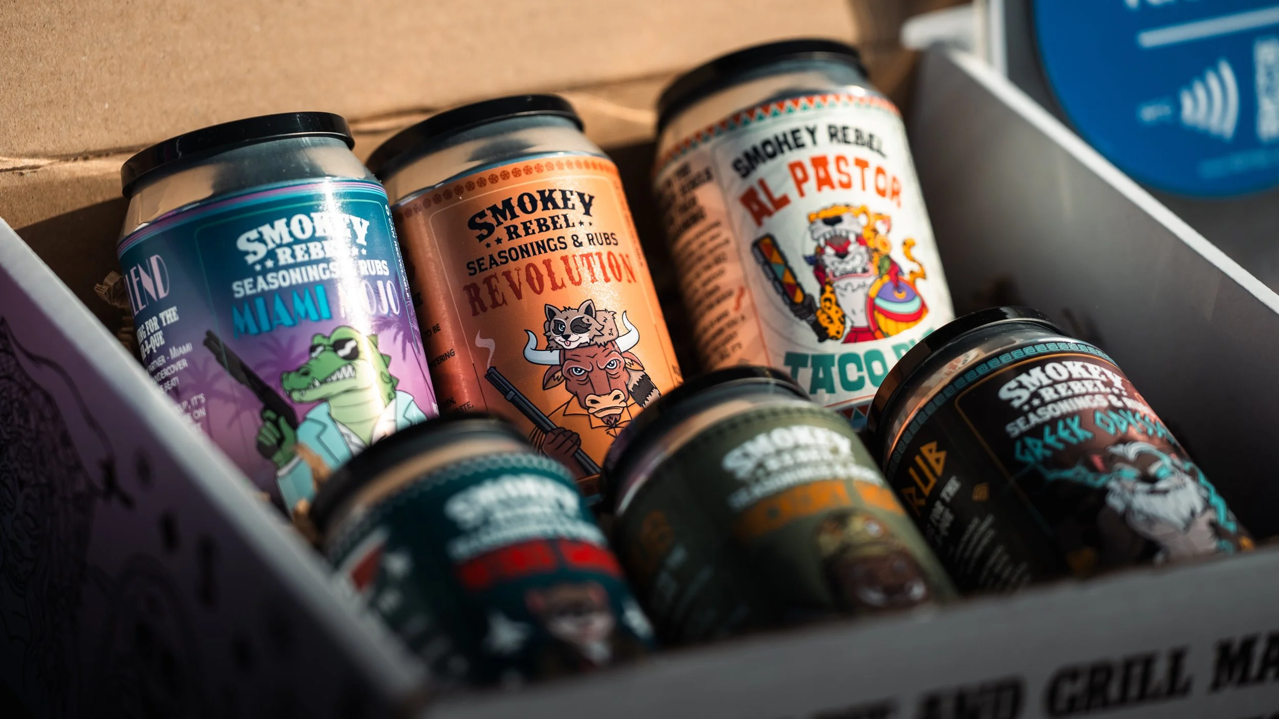 Several jars of Smokey Rebel seasoning and rubs in a box, with colorful cowboy-themed labels and cartoon illustrations of animals.