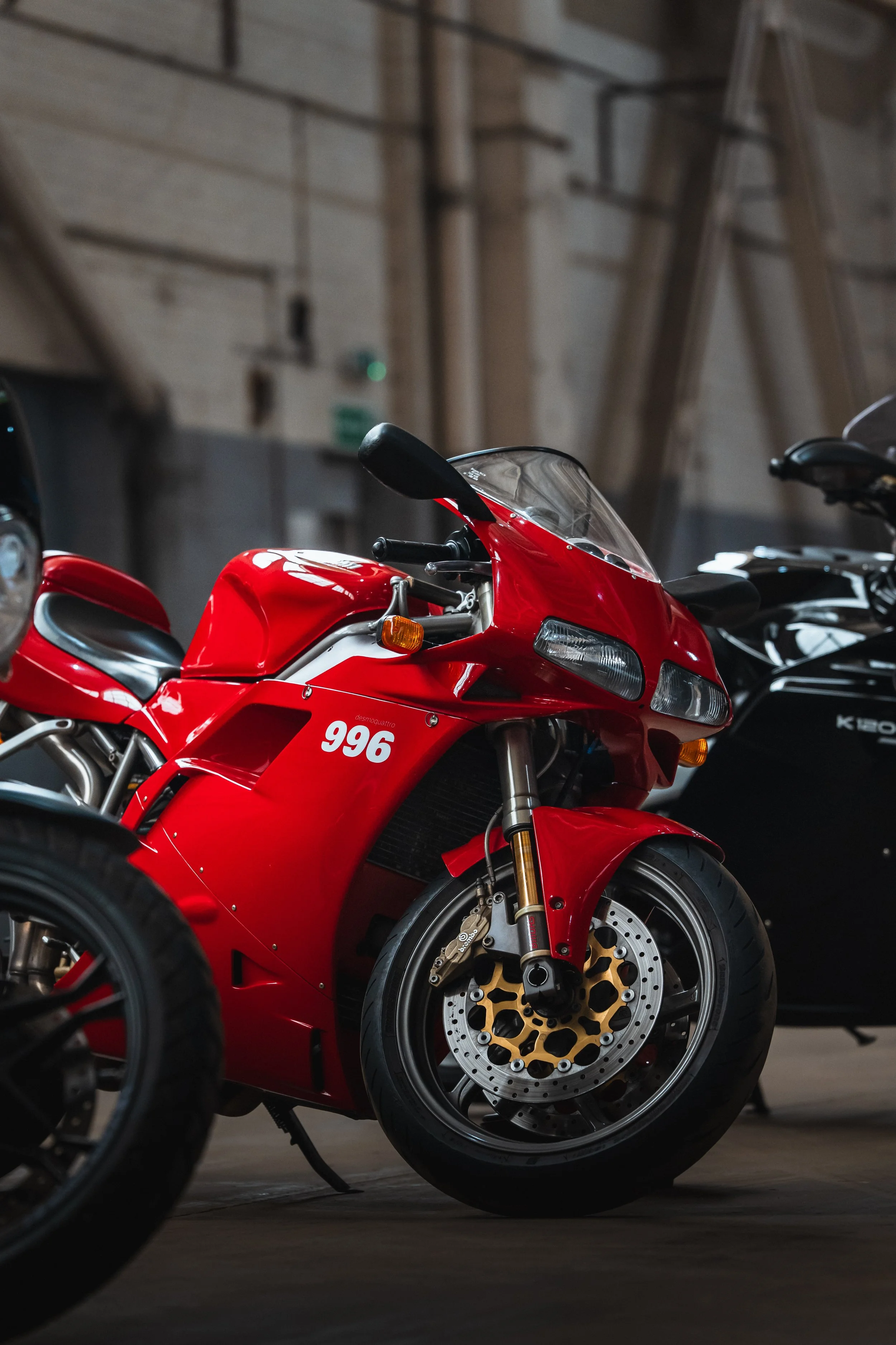 Red sports motorcycle with gold and black accents parked in a dimly lit indoor garage, with a black motorcycle partially visible beside it.